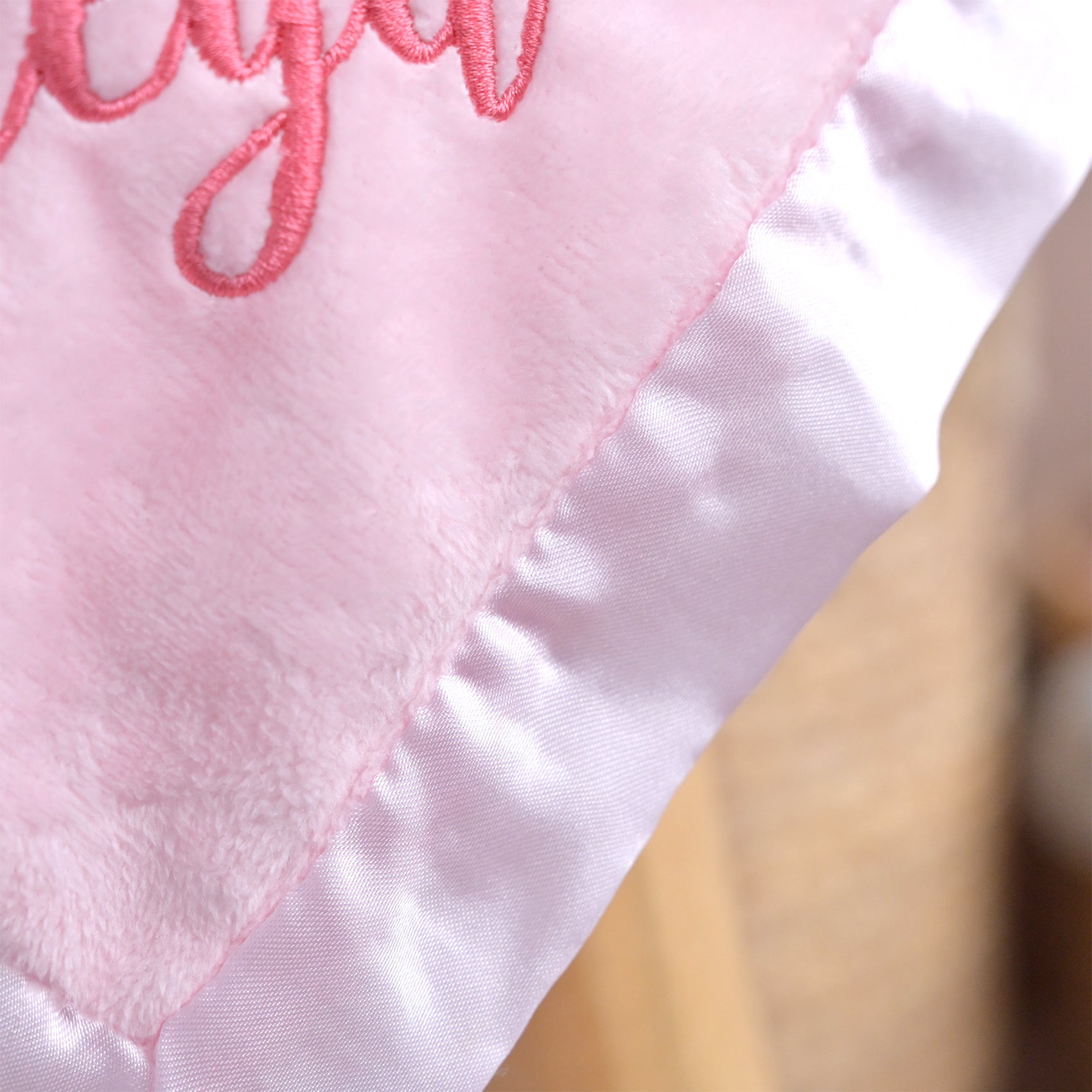 Close-up of a light pink fabric item with embroidered script lettering and a satin ruffled trim, shown against a softly blurred warm-toned background.