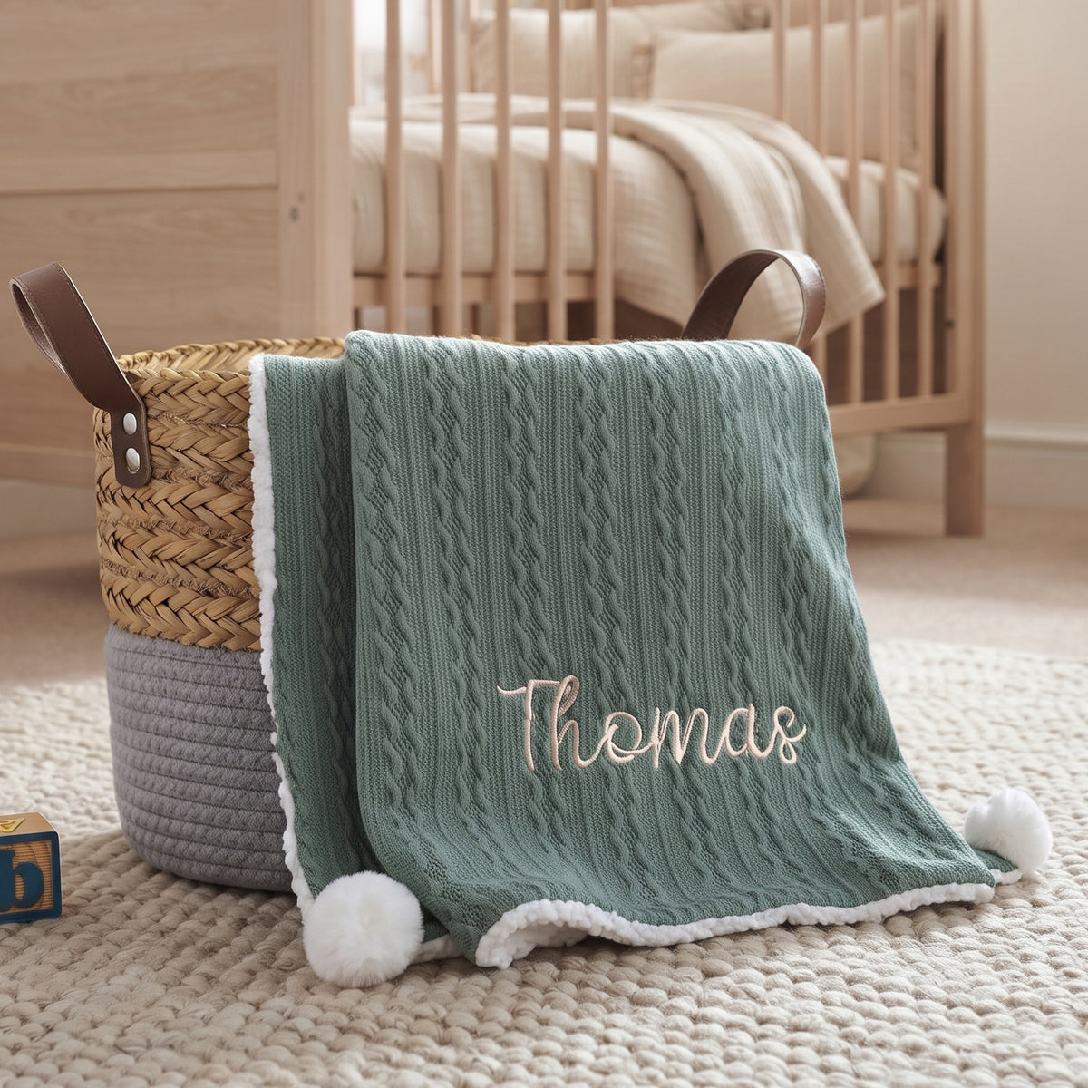 A personalized baby blanket in sage green cable knit with white pom-poms and a white sherpa trim, embroidered with the name 'Thomas' in light pink. The blanket is draped over a two-tone woven basket in a nursery setting.