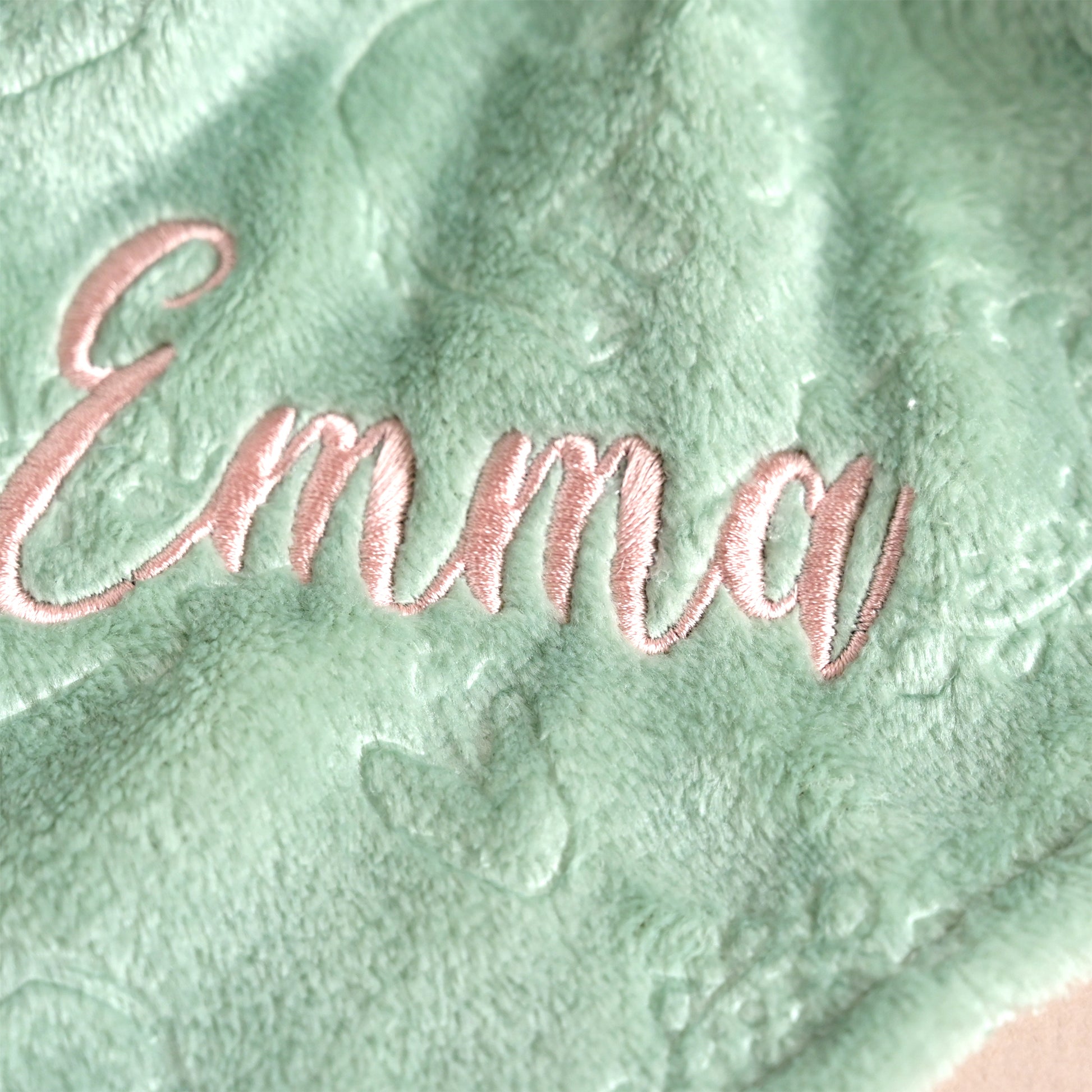 A close-up of a mint green plush blanket with the name 'Emma' embroidered in rose gold script across the front.