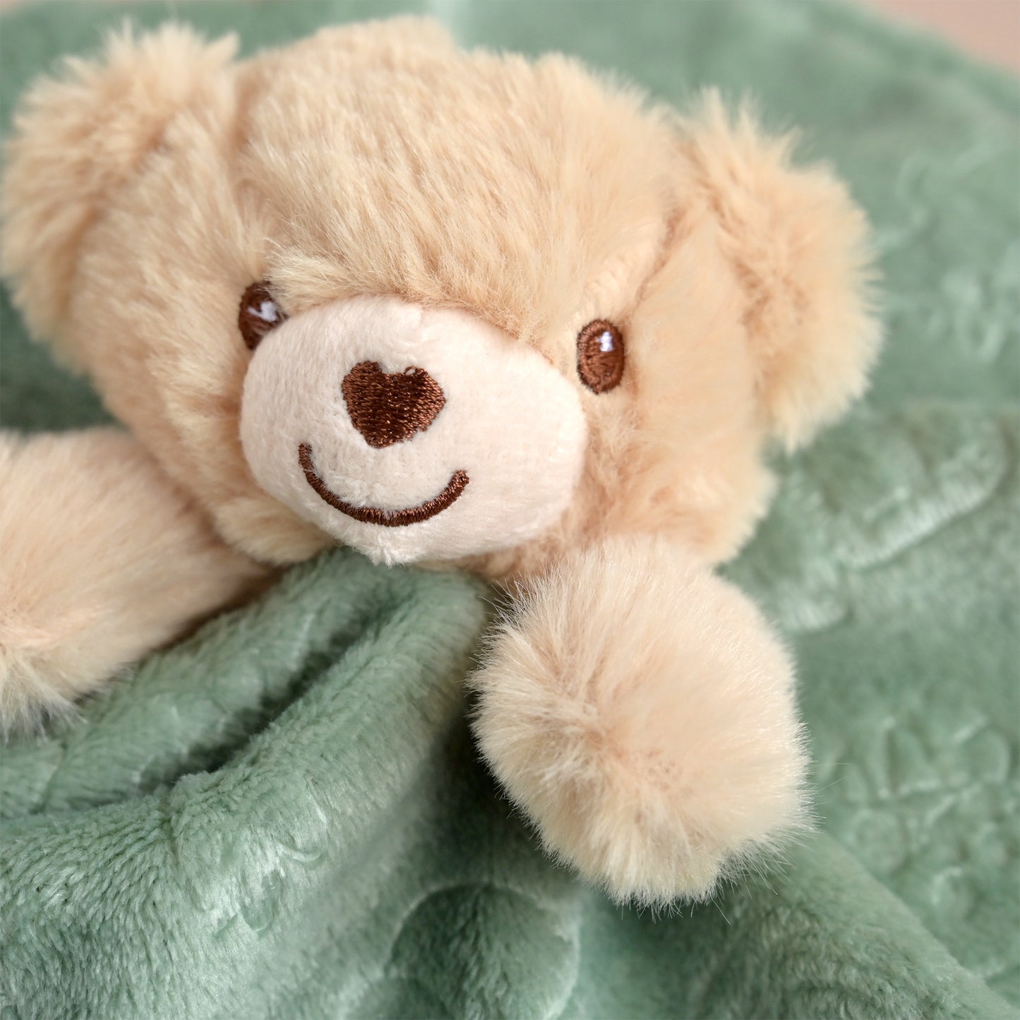 A close-up of a beige plush teddy bear with a white snout and embroidered brown smile, resting on a soft green blanket.