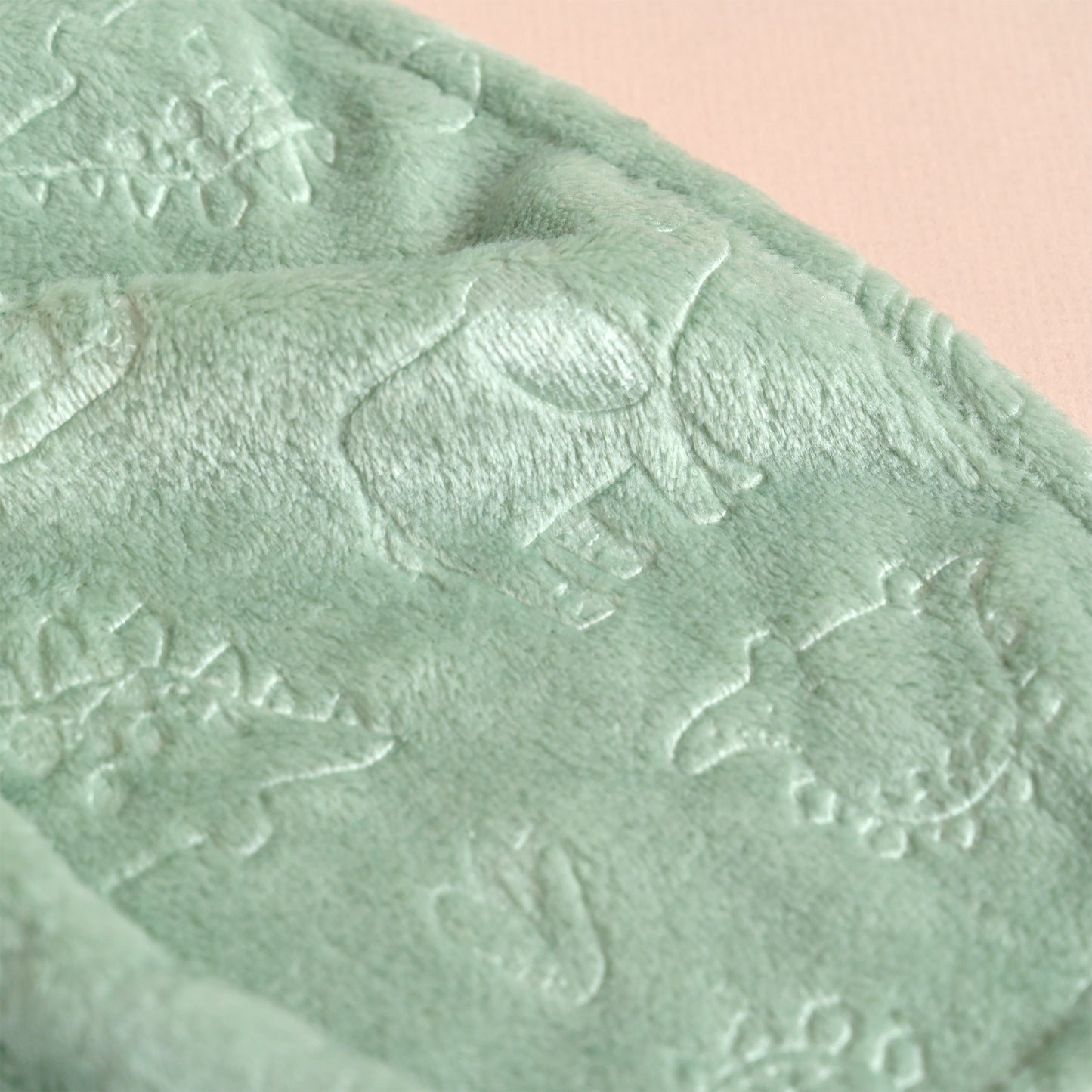 A close-up of a soft, plush sage green blanket featuring embossed animal and floral patterns, set against a solid pale pink background.