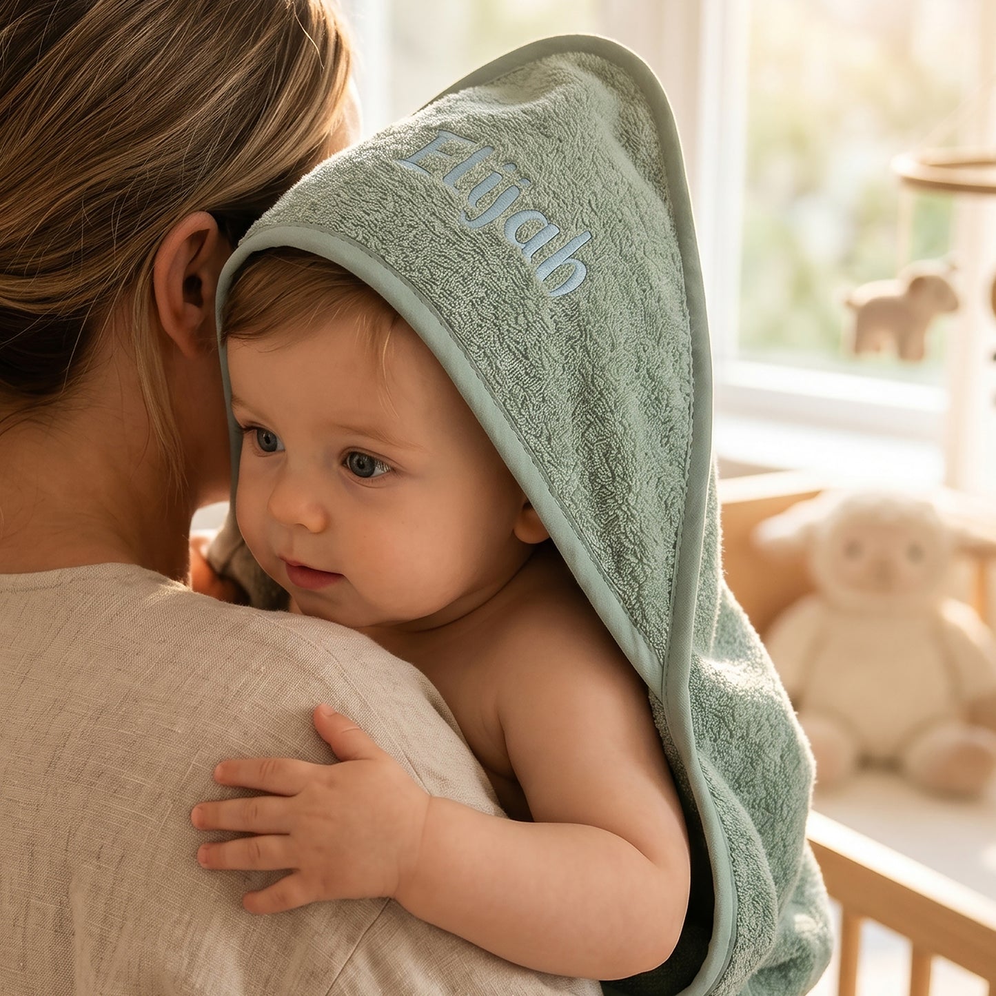 Sage green hooded baby towel in cotton terry, personalised with the name Elijah embroidered in pale blue on the hood, worn by a baby being held over a shoulder.
