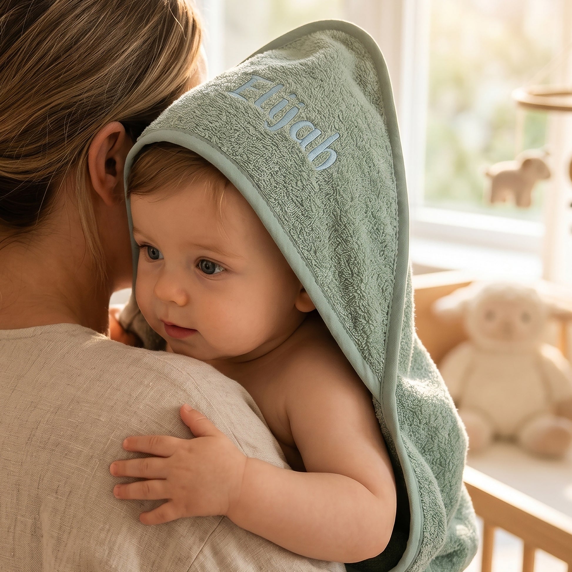 Sage green hooded baby towel in cotton terry, personalised with the name Elijah embroidered in pale blue on the hood, worn by a baby being held over a shoulder.