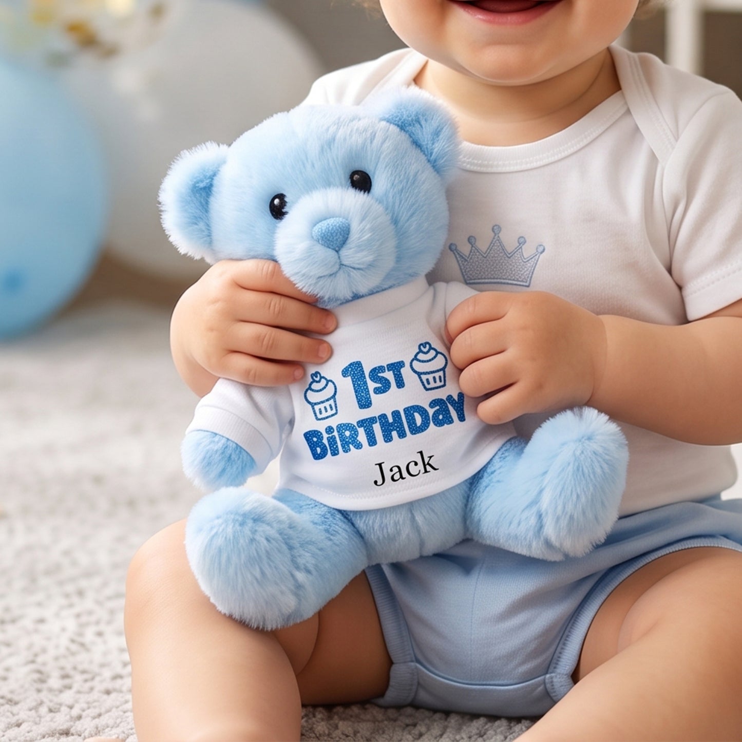 A baby wearing a white onesie with a blue crown graphic sits on a carpeted floor holding a plush light blue teddy bear. The teddy bear wears a white shirt printed with '1st Birthday' and the name 'Jack' in blue.