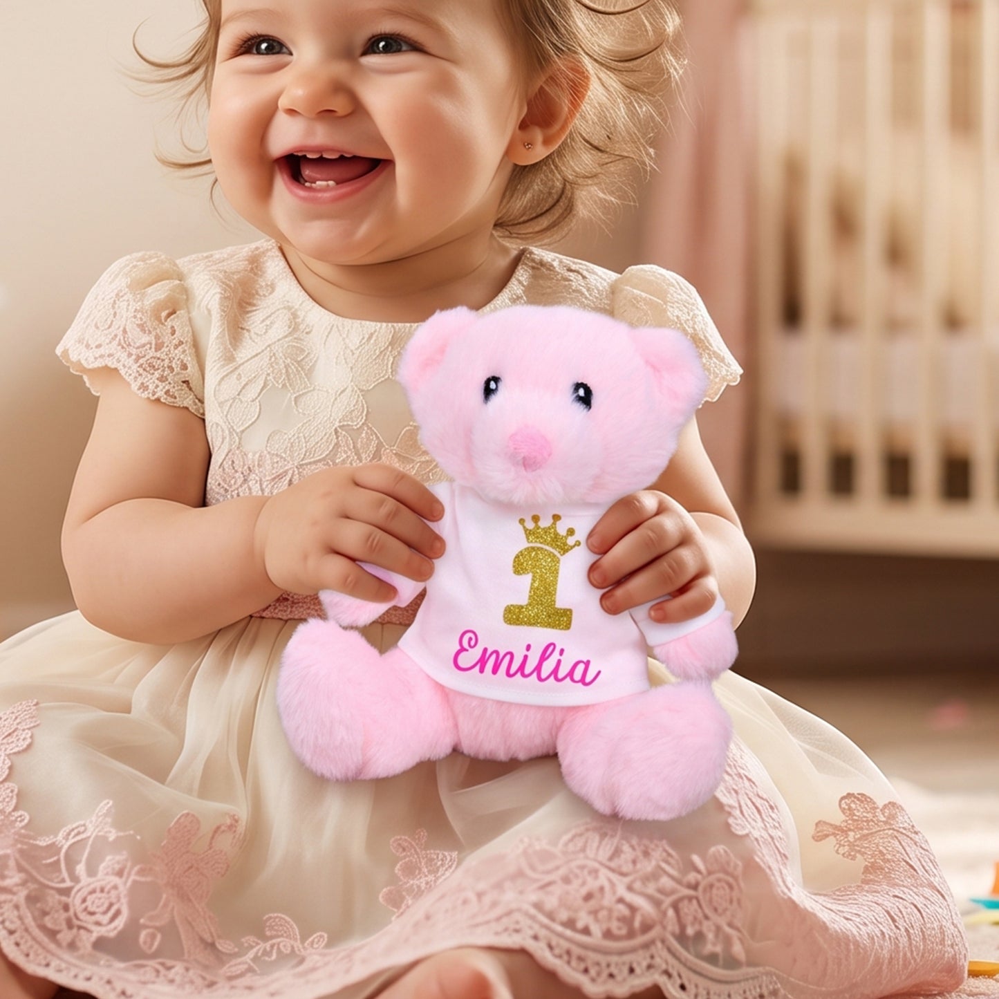 A smiling toddler in a cream lace dress holds a pink plush teddy bear wearing a white bib with a gold number 1 and the name 'Emilia' in pink script, set in a warm indoor room.