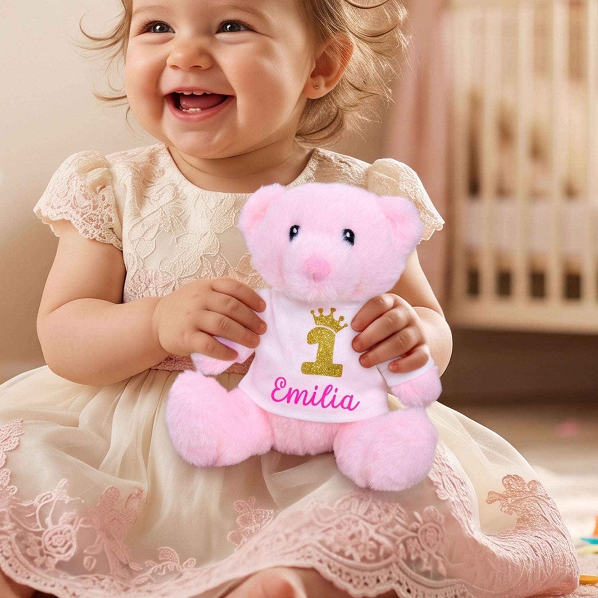 A smiling toddler in a cream lace dress holds a pink plush teddy bear wearing a white bib with a gold number 1 and the name 'Emilia' in pink script, set in a warm indoor room.