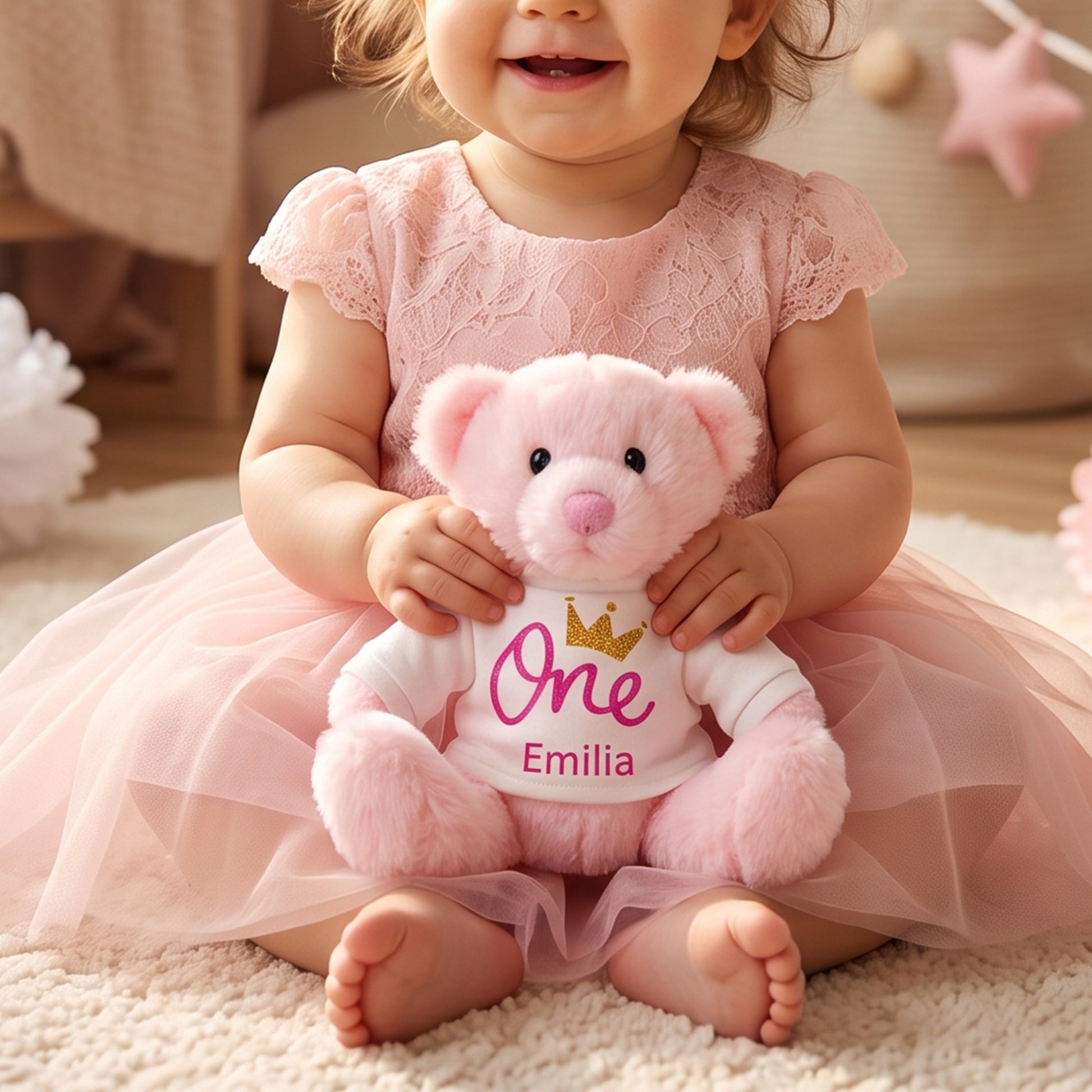 A baby girl in a pink lace dress sits on a plush rug holding a pink teddy bear wearing a white 'One Emilia' t-shirt with a gold crown.