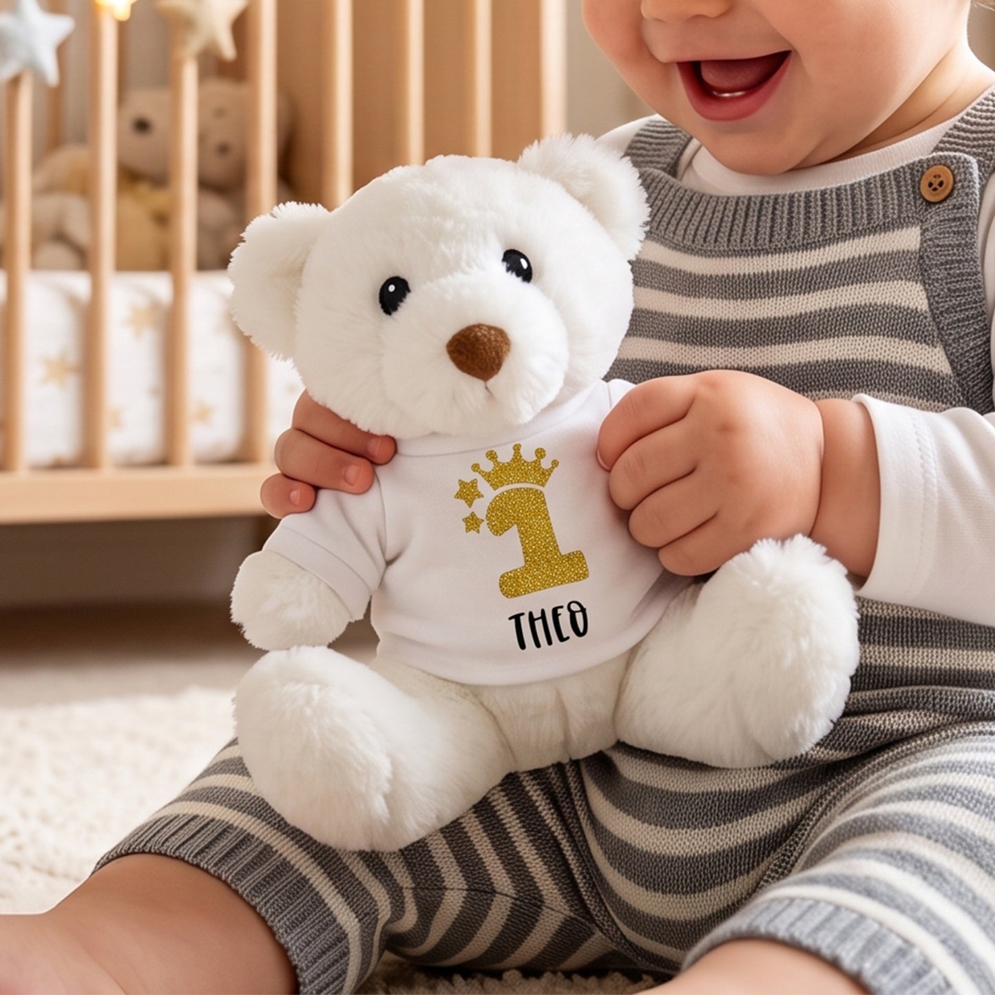A baby in gray and white striped overalls holds a white plush teddy bear wearing a custom white shirt with a gold glittery number 1, stars, crown, and the name 'THCO'.