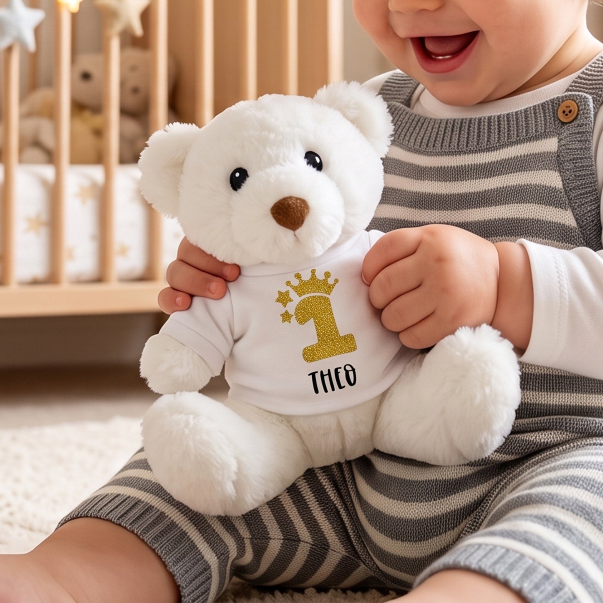 A baby in gray and white striped overalls holds a white plush teddy bear wearing a custom white shirt with a gold glittery number 1, stars, crown, and the name 'THCO'.