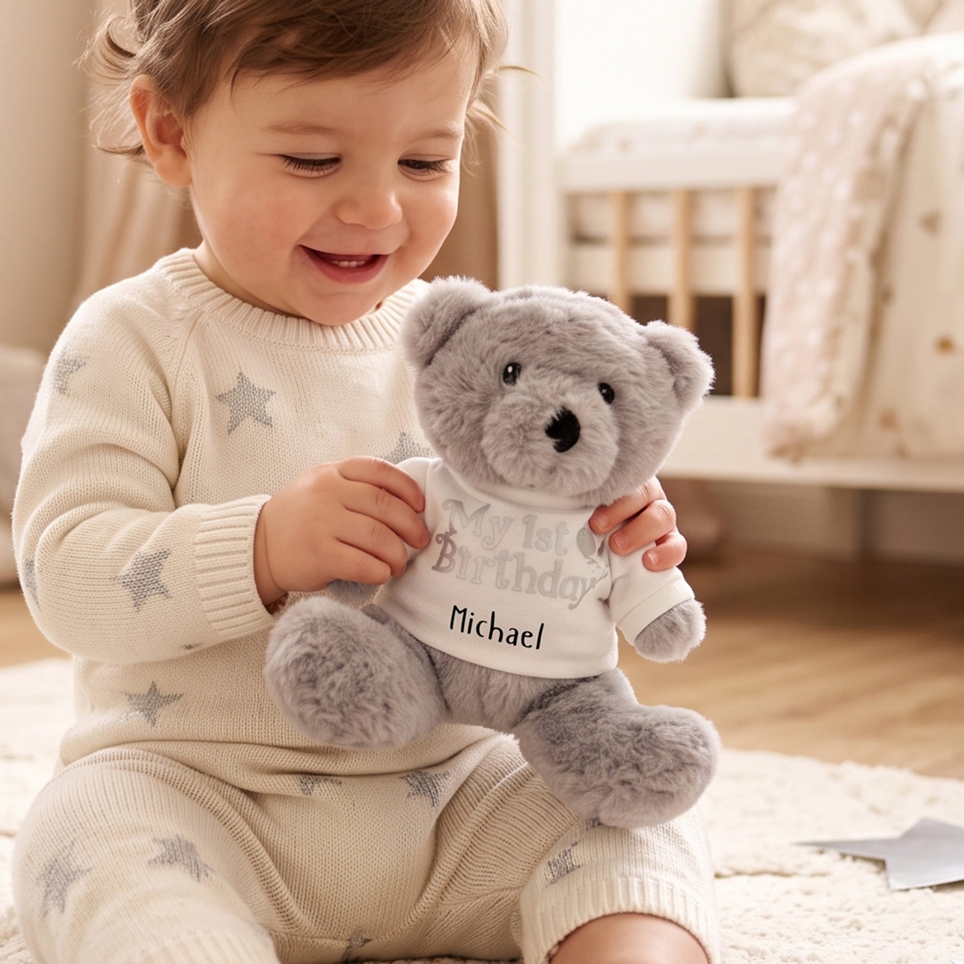 Grey teddy bear soft toy wearing a white t-shirt printed with "My 1st Birthday" and a balloon design, personalised with the name Michael, held by a toddler.