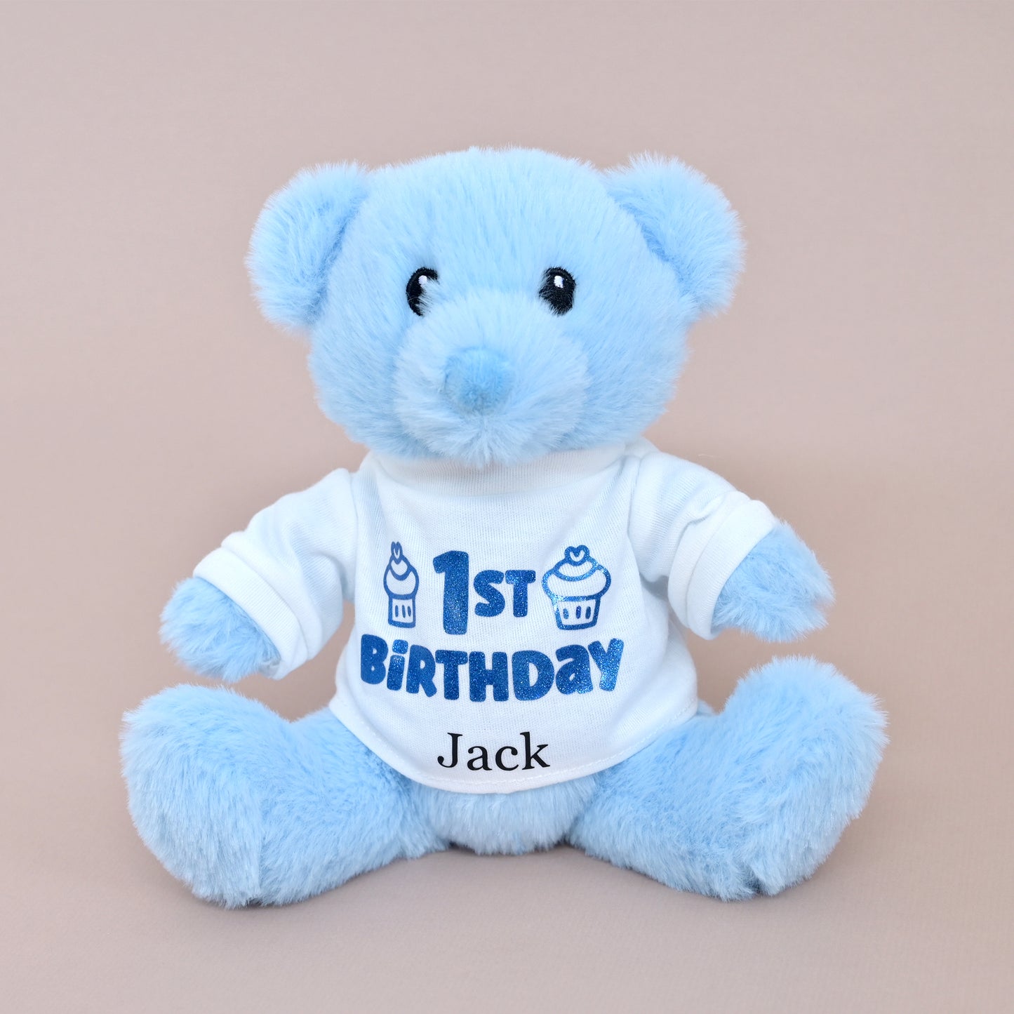 A light blue plush teddy bear wearing a white short-sleeve shirt with blue '1st Birthday' text and cupcake graphics, plus the name 'Jack' printed below, photographed against a plain warm beige background.