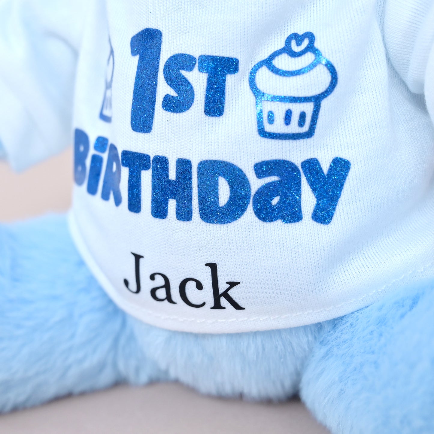 Close-up of a plush toy or stuffed animal wearing a white shirt with blue glitter text reading '1st Birthday' and a cupcake graphic, plus the name 'Jack' in black below.