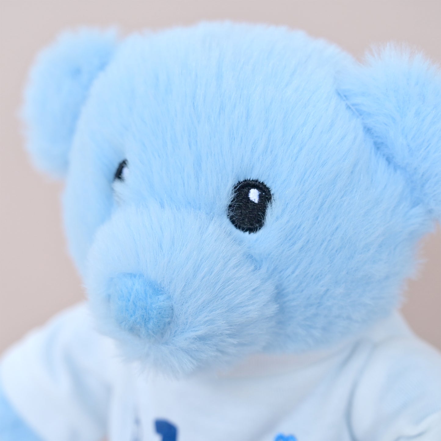 A close-up of a light blue plush teddy bear wearing a white shirt with small blue heart details, set against a soft beige background.