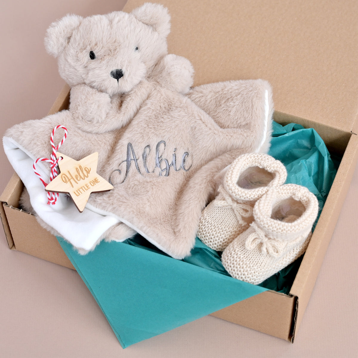 A personalized baby gift set in a cardboard box, featuring a plush teddy bear security blanket with 'Albie' embroidered on it, a white baby towel with a wooden star tag that reads 'Hello little star', and a pair of cream knit baby booties on a teal tissue paper background.