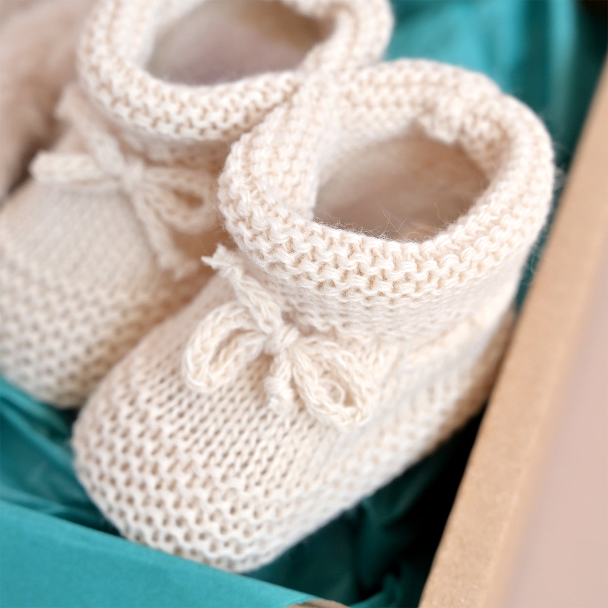 A pair of cream-colored hand-knit baby booties with small bows, nestled in a box lined with teal paper.