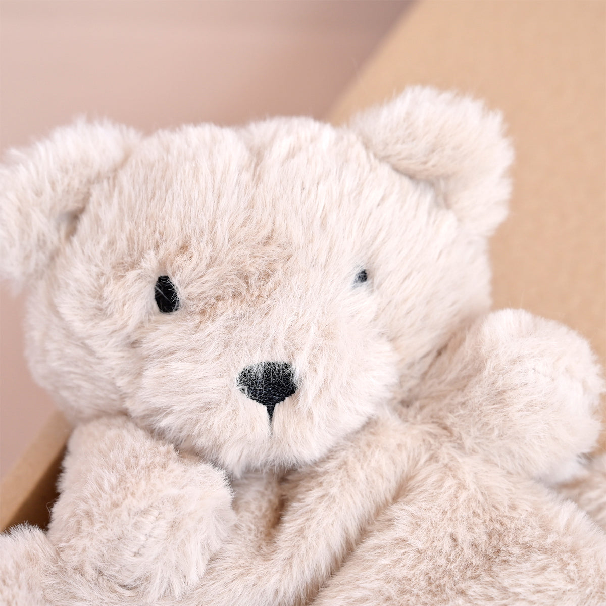 A close-up of a soft, cream-colored plush teddy bear with small black embroidered eyes and nose, resting against a neutral tan background.
