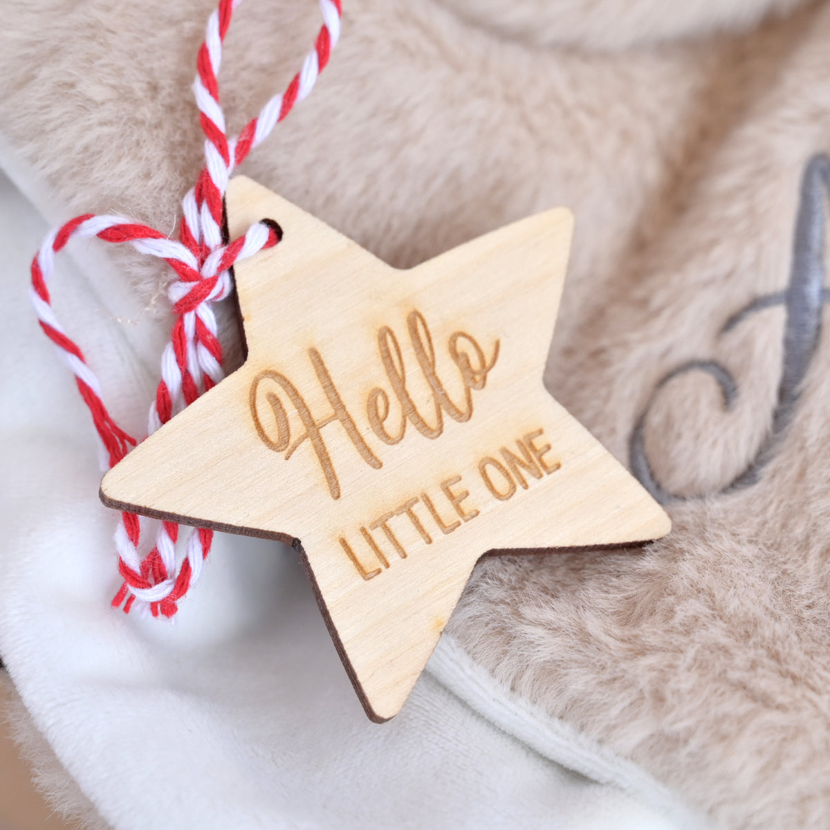 A star-shaped wooden ornament engraved with 'Hello LITTLE ONE' in a warm natural wood tone, hanging from a red-and-white twisted string on soft beige faux fur fabric.