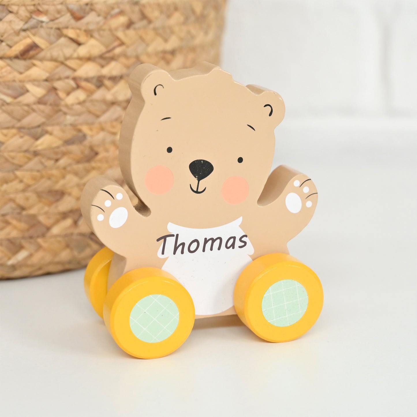 A personalized wooden bear toy with yellow wheels and a white belly patch that reads 'Thomas', photographed on a light surface with a woven basket in the background.
