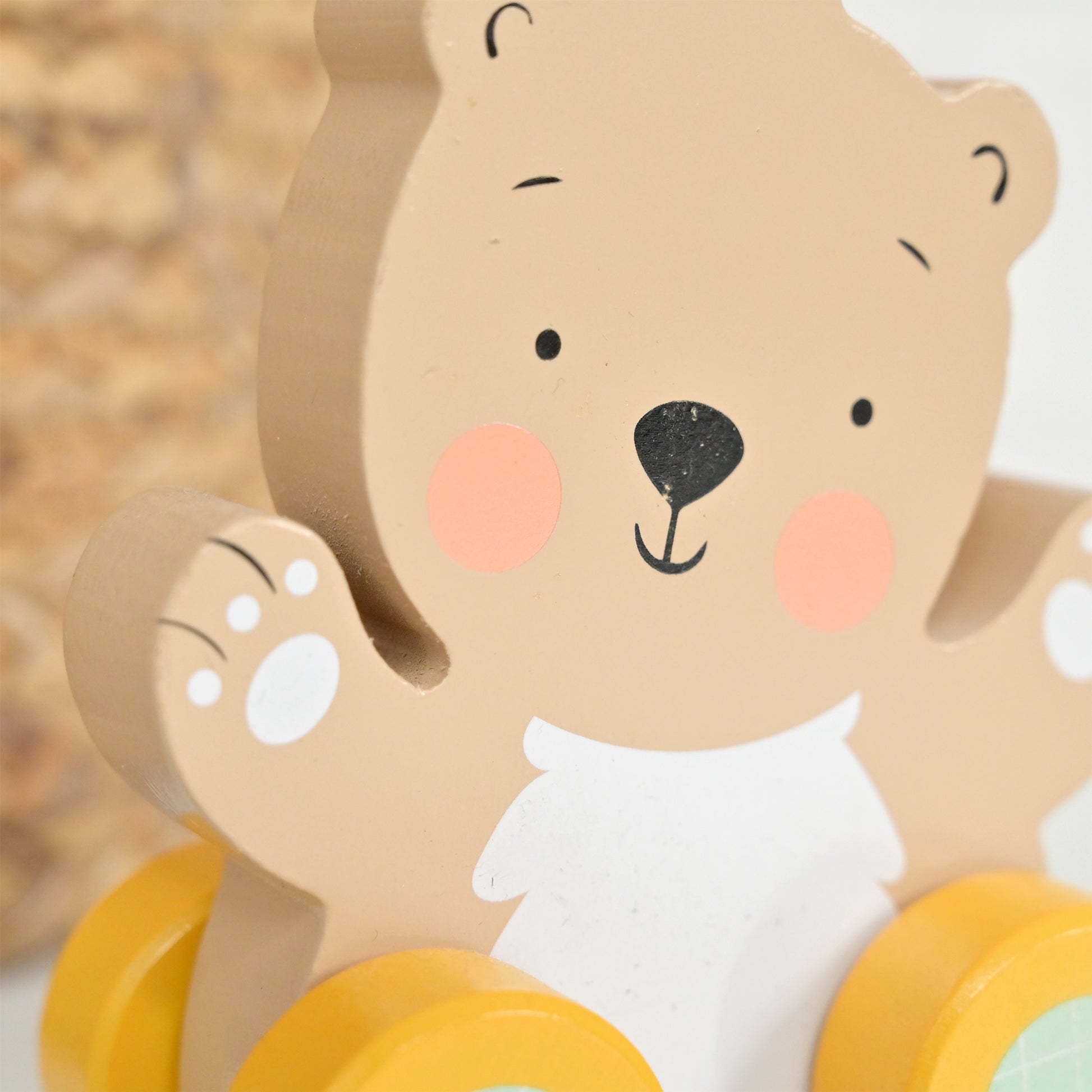 Close-up of a wooden bear toy with a beige body, black facial features, pink cheeks, and yellow wheels at the bottom.