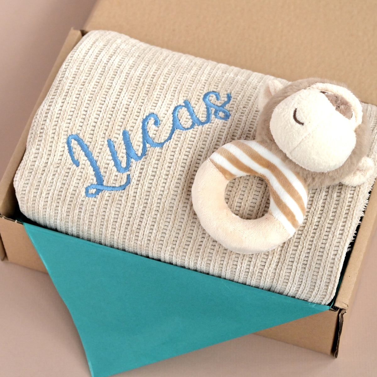 A personalized baby blanket gift set in a box, featuring a cream ribbed knit blanket with the name 'Lucas' embroidered in blue, a plush monkey toy, and a teal tissue paper lining inside a brown cardboard box.