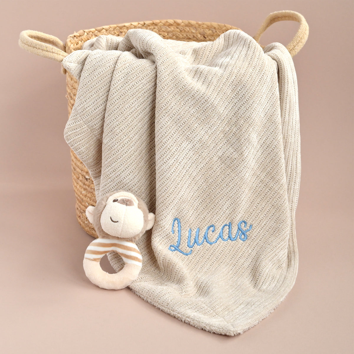 A cream-colored textured baby blanket with blue embroidered name 'Lucas' draped over a woven straw basket, styled with a plush monkey teether toy on a solid dusty pink background.