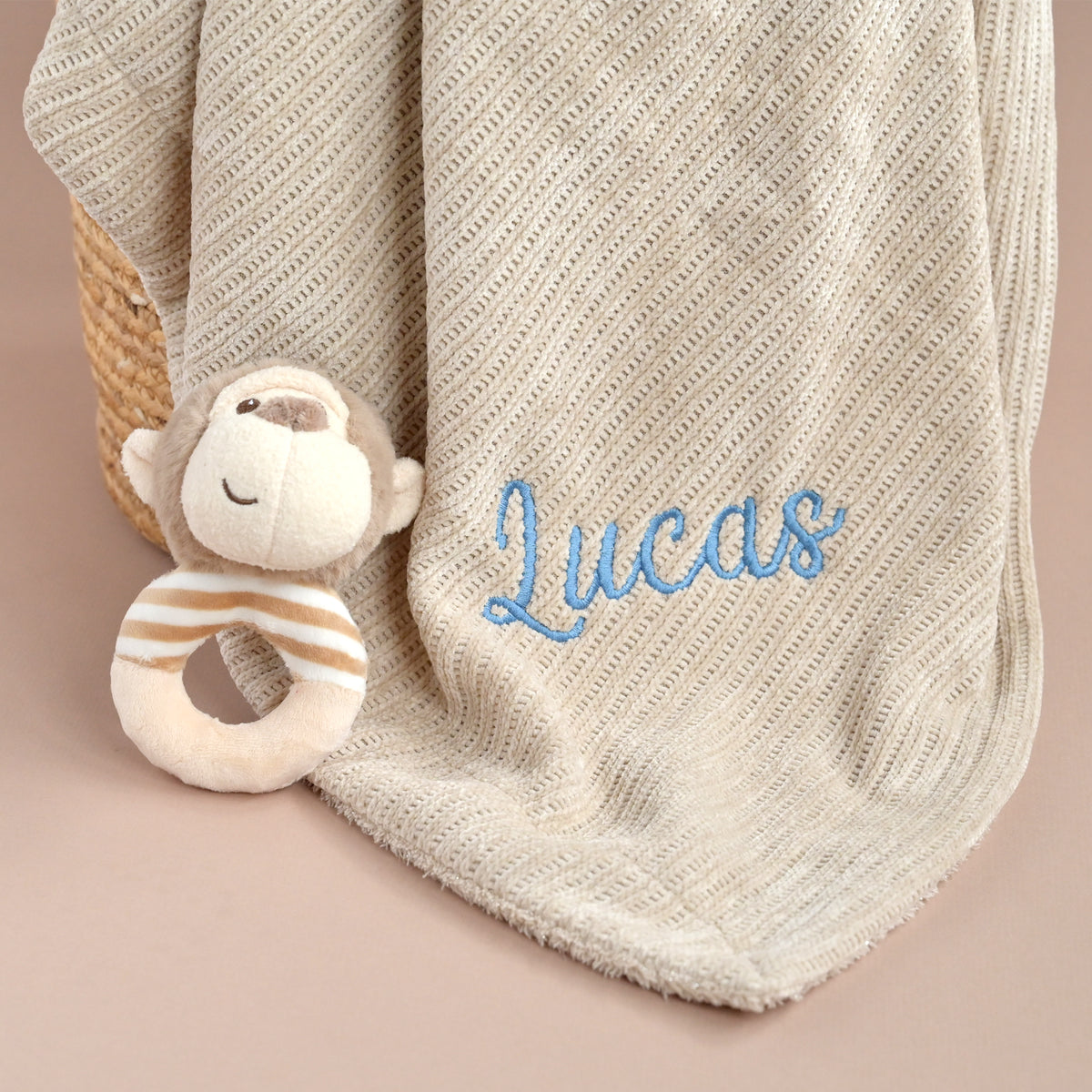 A personalized baby blanket in cream with diagonal knit texture, featuring the name 'Lucas' embroidered in blue script. A plush monkey teething ring toy is tucked under the corner of the blanket, which is draped over a woven basket against a solid beige background.