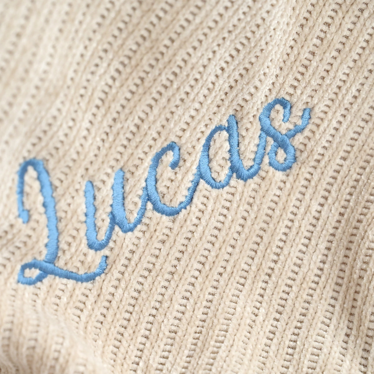 Close-up of a beige ribbed knit fabric with light blue embroidered script reading 'Lucas'.