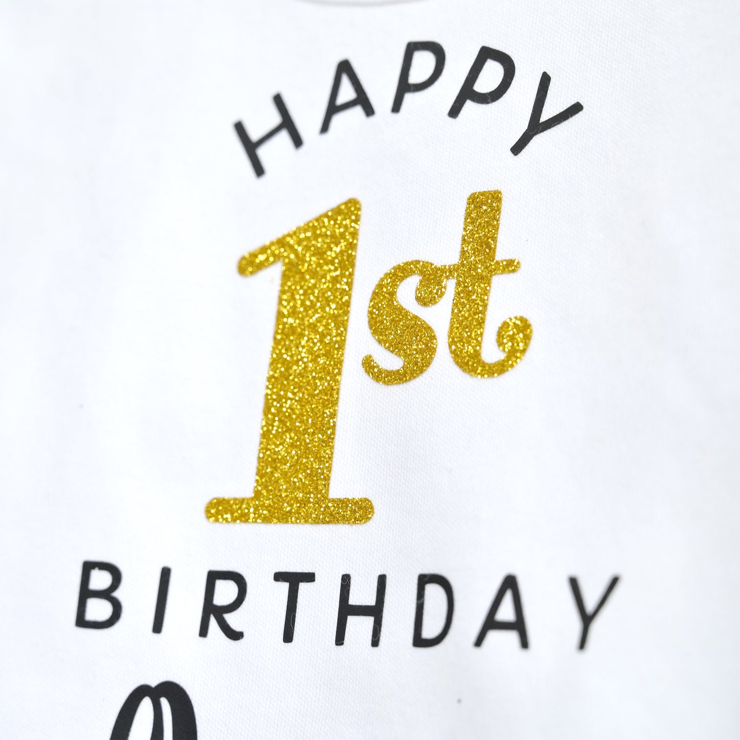 Close-up of a white garment printed with 'HAPPY 1st BIRTHDAY' in black and glittery gold lettering.