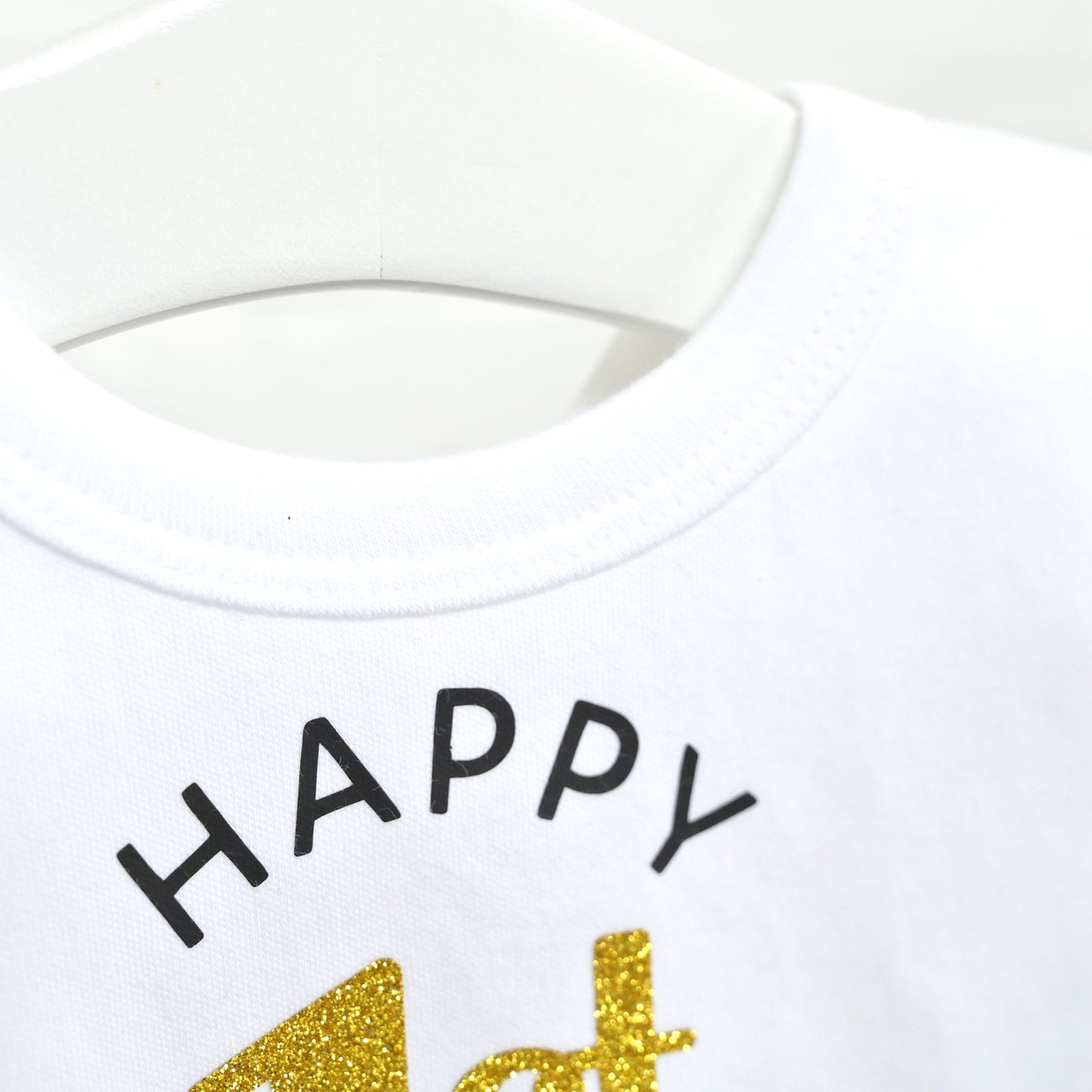 Close-up of a white crewneck shirt on a white hanger, featuring the word 'HAPPY' in black arched lettering and a gold glittery '4th' graphic below.