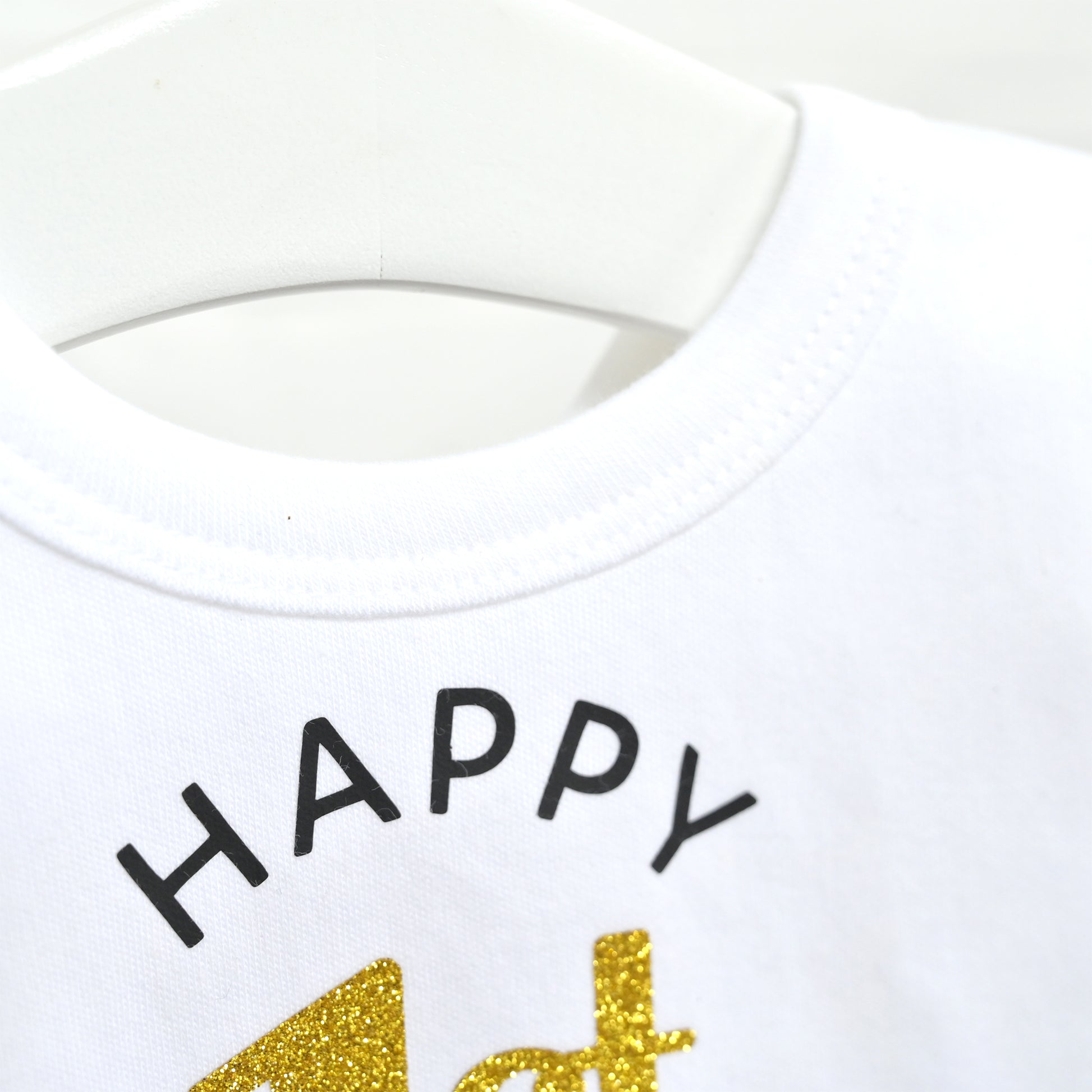 Close-up of a white crewneck shirt on a white hanger, featuring the word 'HAPPY' in black arched lettering and a gold glittery '4th' graphic below.