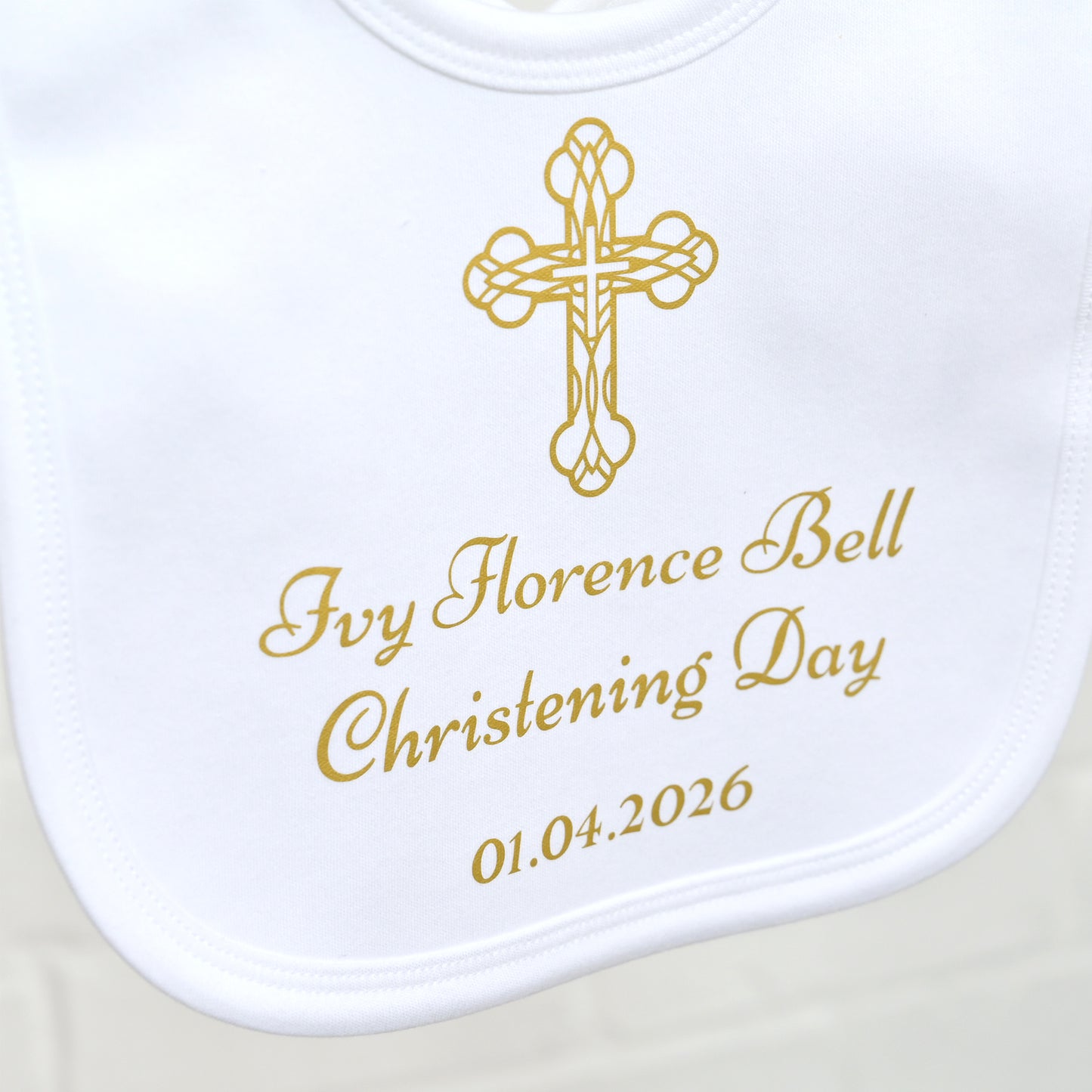 A close-up of a white baby bib with a gold cross and personalized christening text reading 'Ivy Florence Bell Christening Day 01.04.