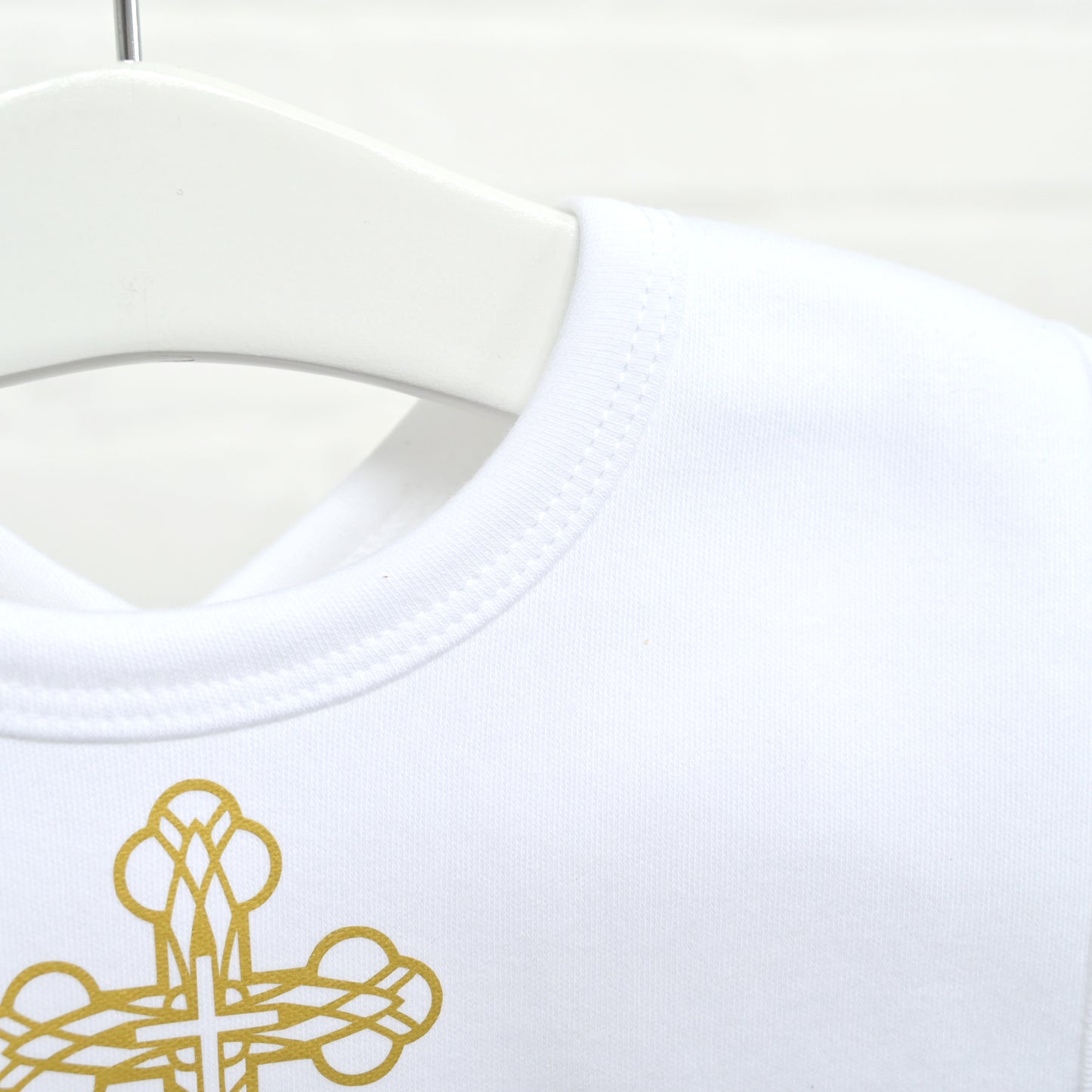 Close-up of a white crew-neck garment on a white hanger, showing a gold embroidered cross motif near the lower left area.