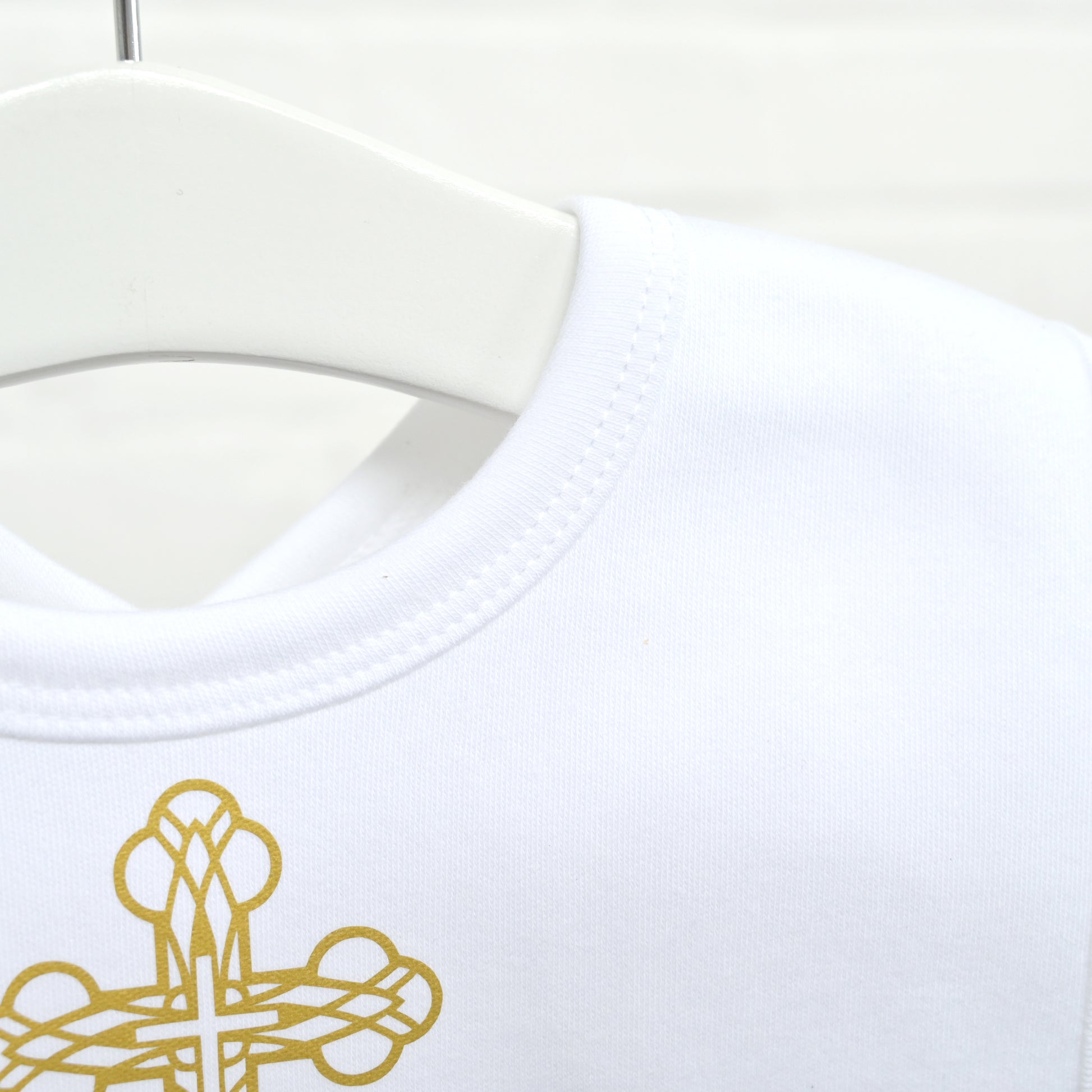 Close-up of a white crew-neck garment on a white hanger, showing a gold embroidered cross motif near the lower left area.