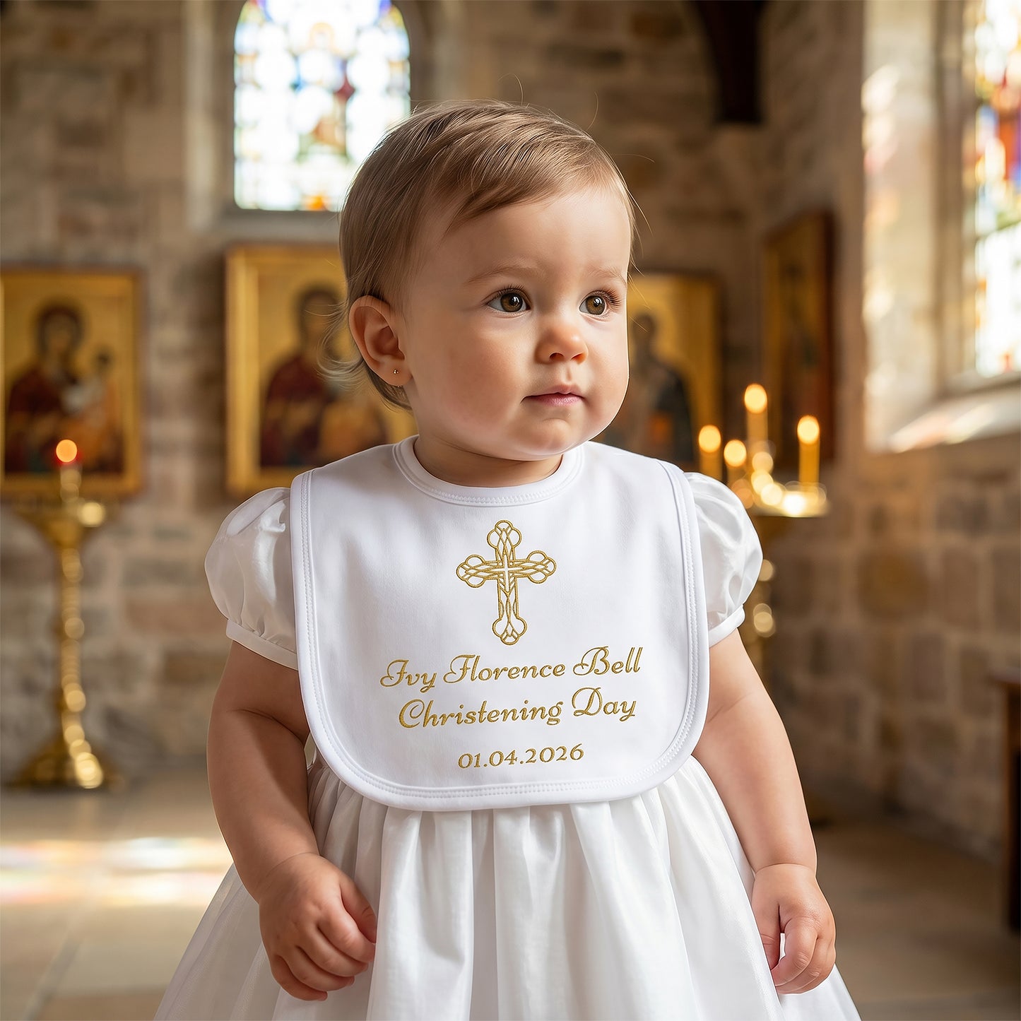 White velcro christening bib printed with a gold Celtic knot cross and personalised with the name Ivy Florence Bell, "Christening Day" and the date 01.04.2026, worn by a baby in a church.