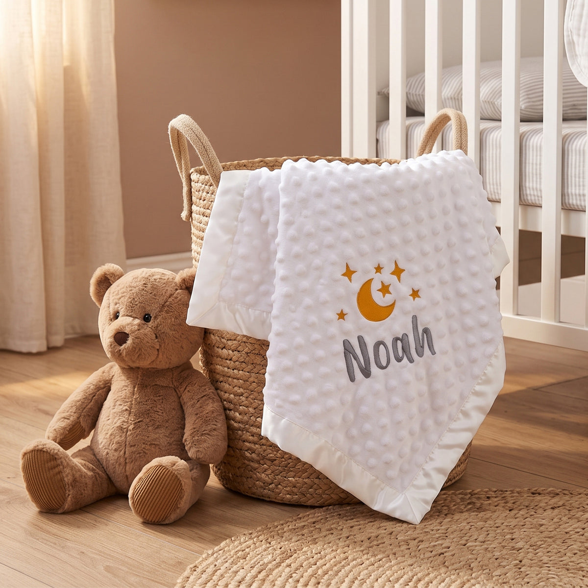 A white textured baby blanket with a custom embroidered name 'Noah' and a crescent moon design, draped over a woven basket next to a brown teddy bear in a nursery setting.