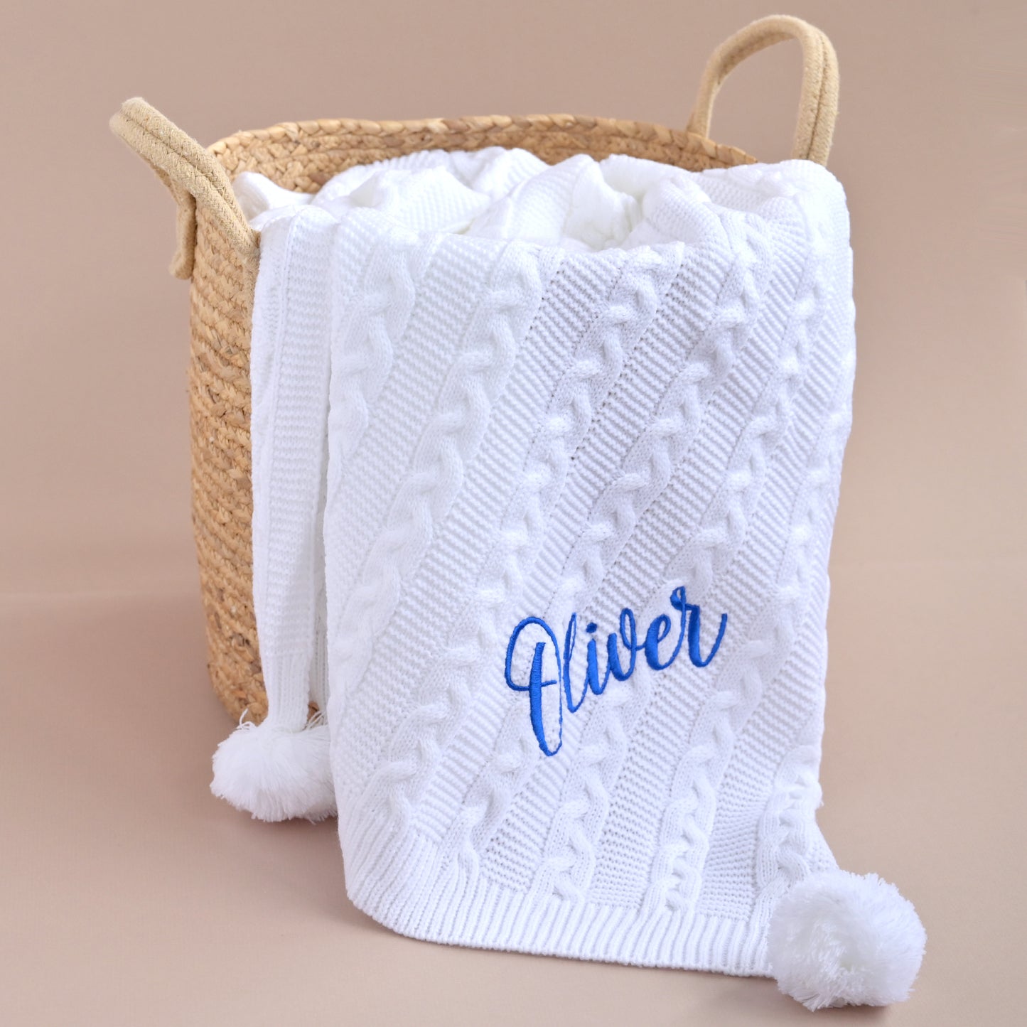 A white cable-knit baby blanket with fluffy pom-poms and blue cursive embroidery reading 'River', draped over a woven straw laundry basket on a warm beige background.