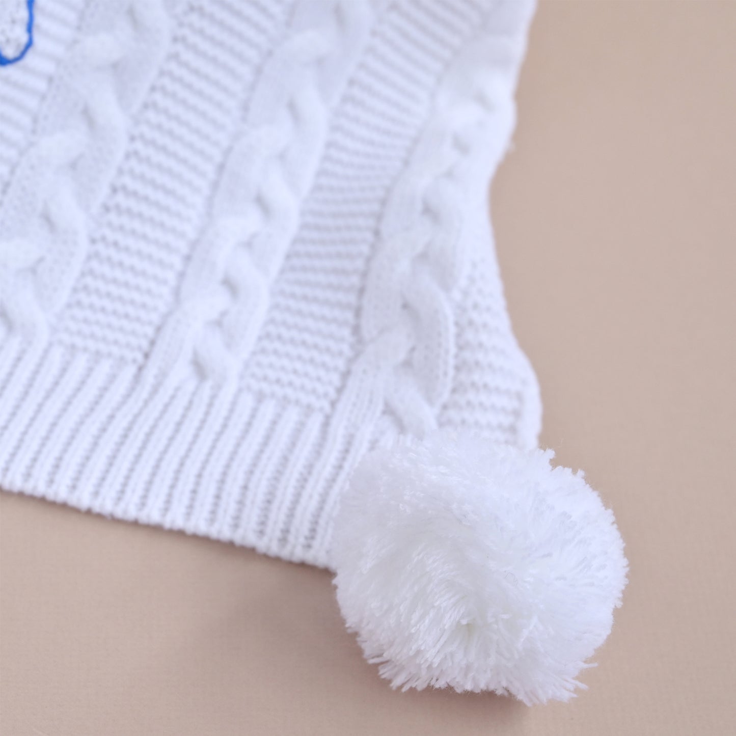 Close-up of a white knit hat with a cable-knit pattern and a fluffy pom-pom, shown on a plain beige background.