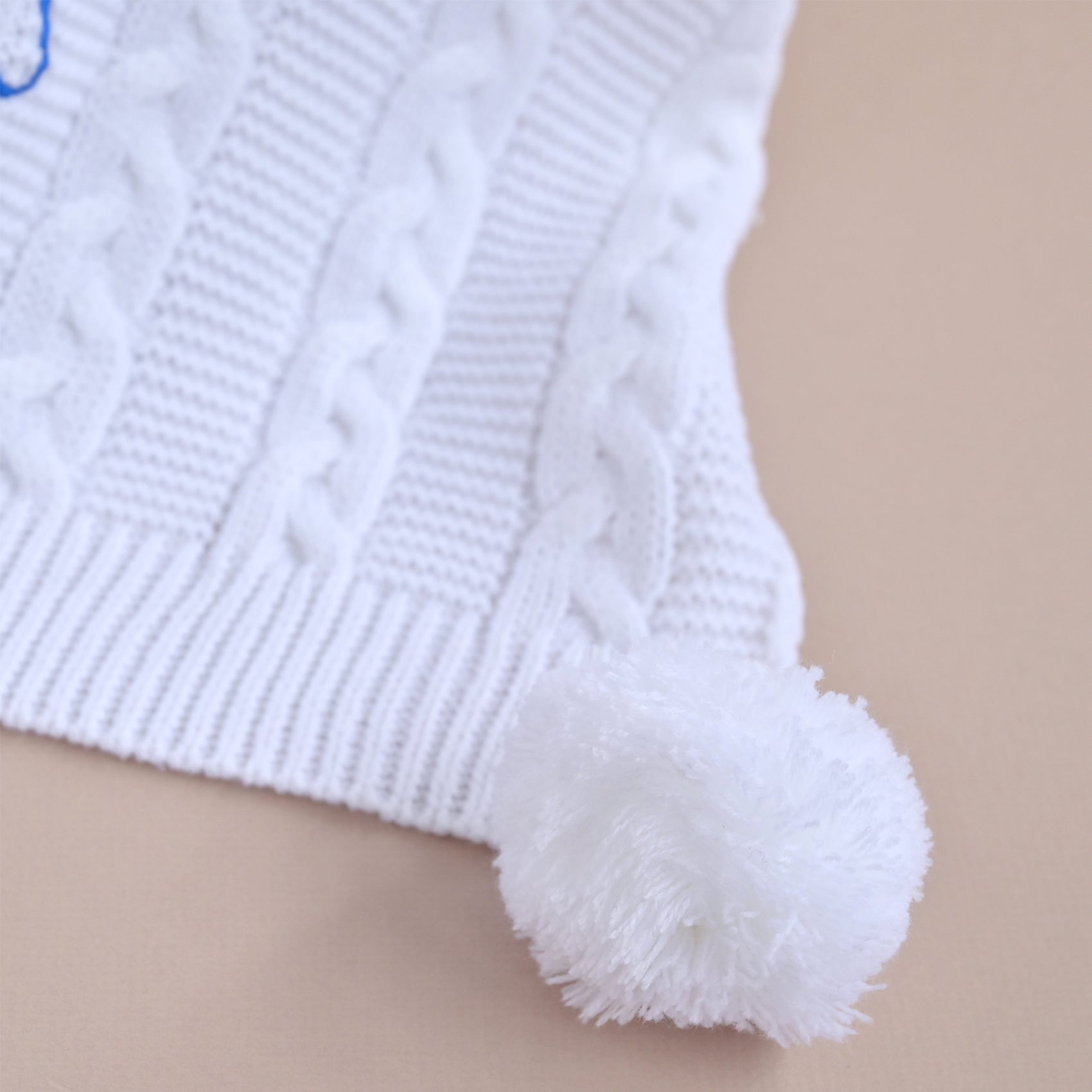 Close-up of a white knit hat with a cable-knit pattern and a fluffy pom-pom, shown on a plain beige background.