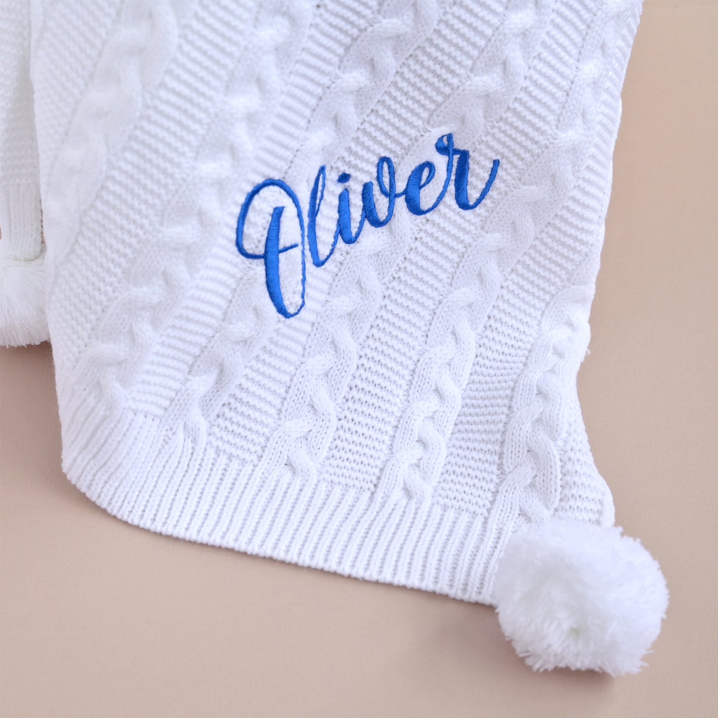 A close-up of a white cable-knit baby blanket with a fluffy pom-pom and personalized blue cursive embroidery reading 'Oliver' on a beige background.