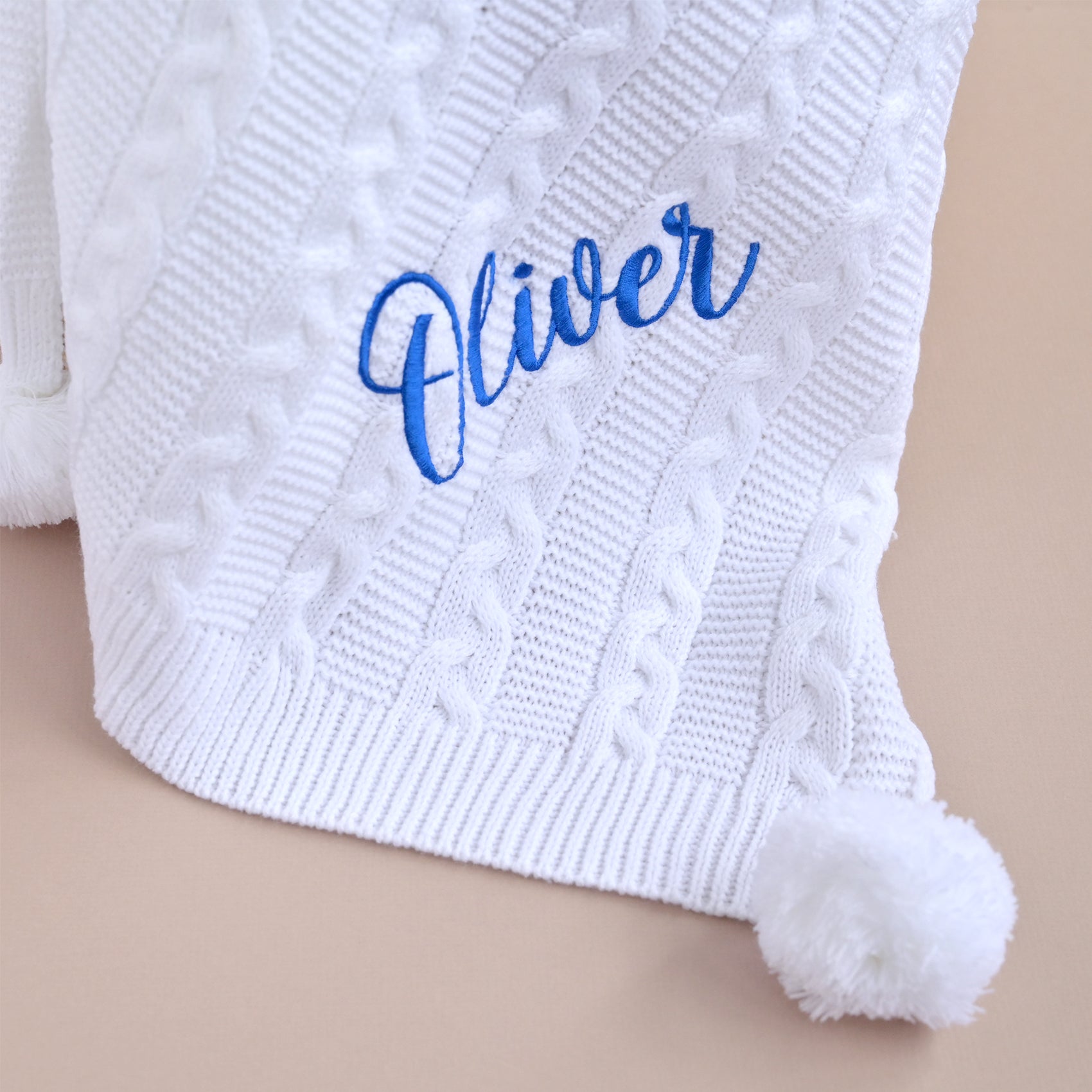 A close-up of a white cable-knit baby blanket with a fluffy pom-pom and personalized blue cursive embroidery reading 'Oliver' on a beige background.
