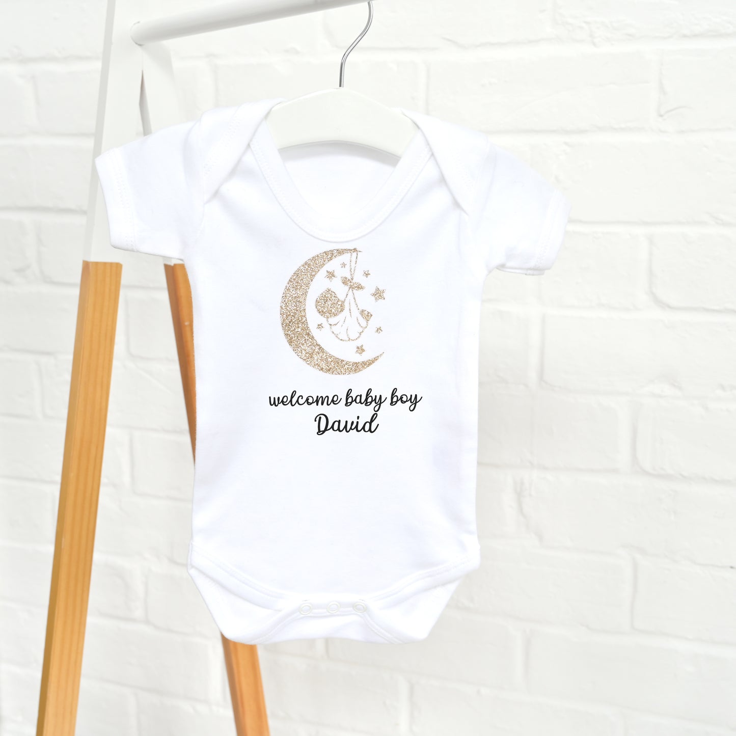 A white short-sleeve baby bodysuit hanging on a wooden hanger against a white brick wall. The bodysuit features a gold glitter crescent moon and stars graphic with a baby bottle hanging from the crescent, and the text 'welcome baby boy David' printed below in black.