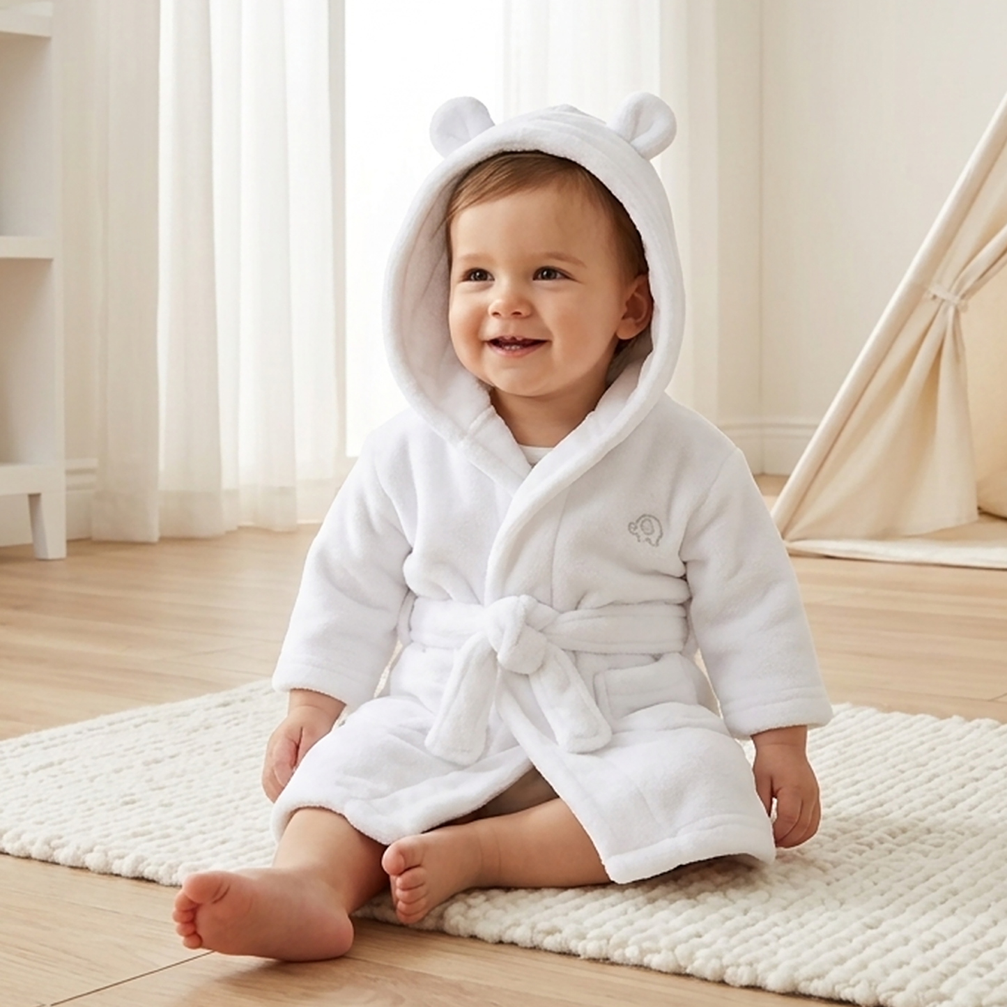 A baby wearing a white plush hooded bathrobe with bear ears sits on a textured cream rug in a bright, neutral-toned room.