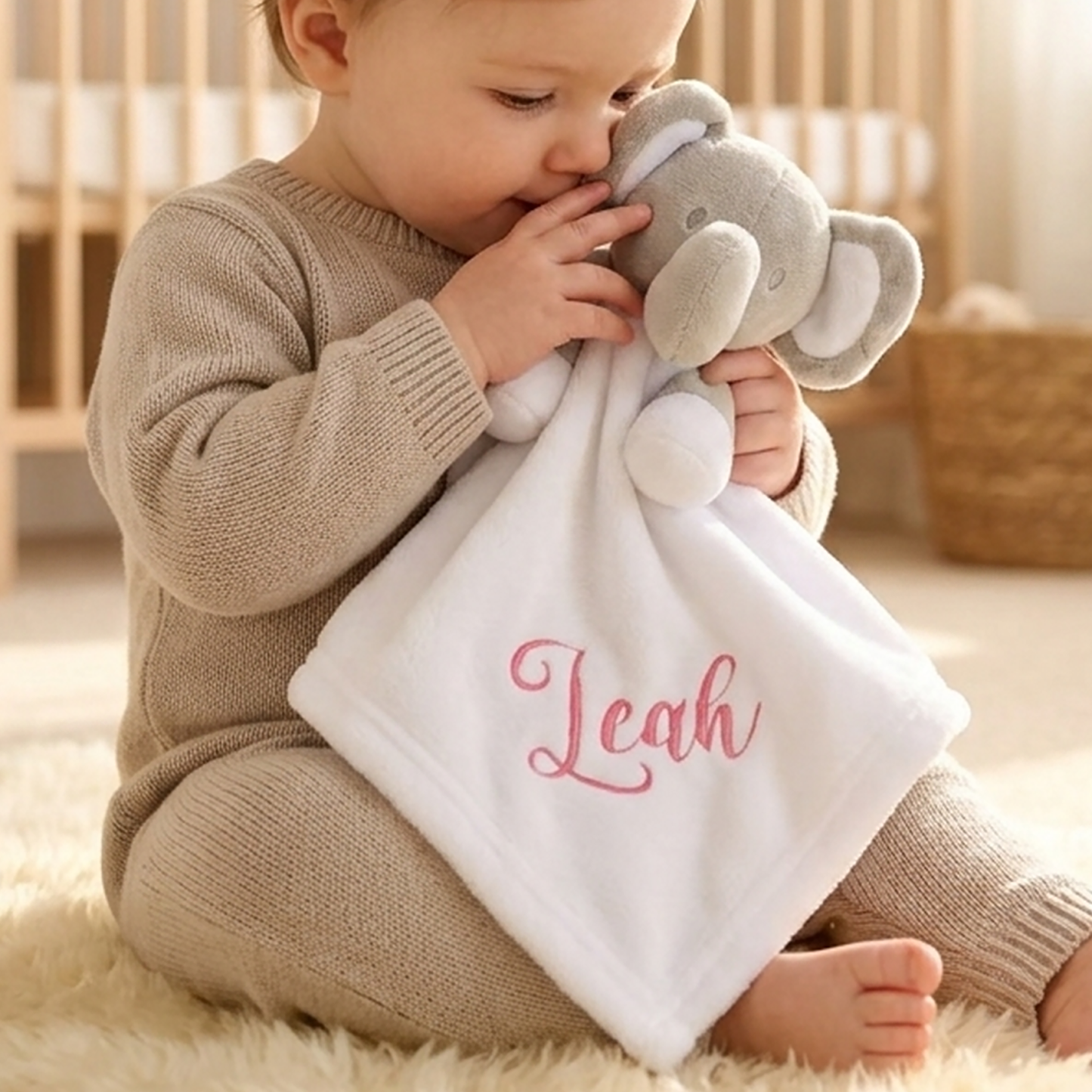 Baby holding a white elephant comforter, personalised with the name "Leah" in pink script embroidery.