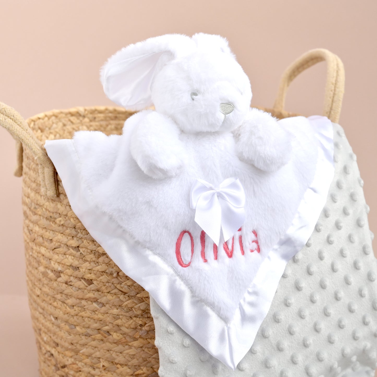 A white plush bunny security blanket with a white bow and the name 'Olivia' embroidered in red, draped over a woven straw basket next to a textured white blanket.