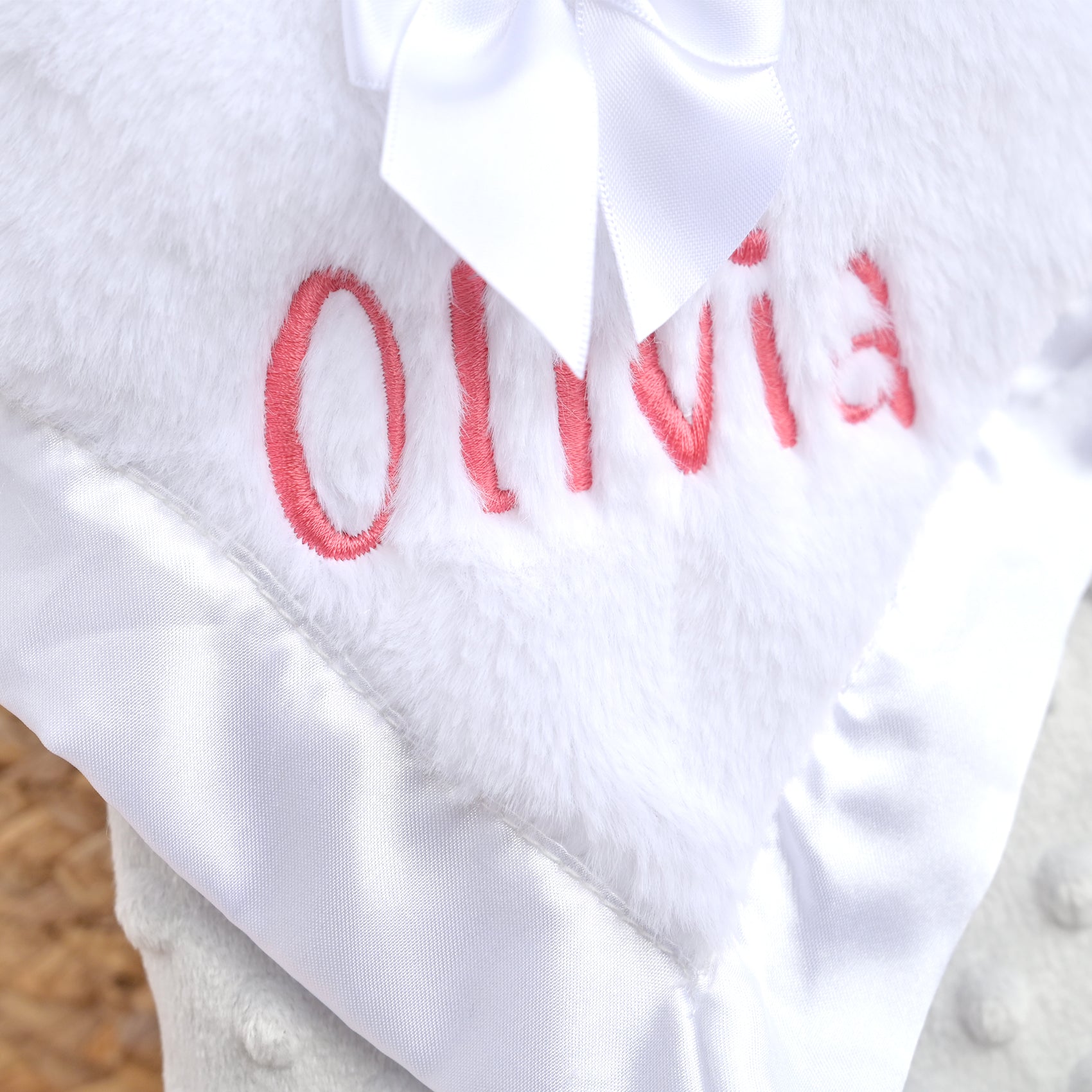 Close-up of a white plush baby blanket with red embroidered name 'Olivia' and a white satin bow, shown against a soft neutral background.
