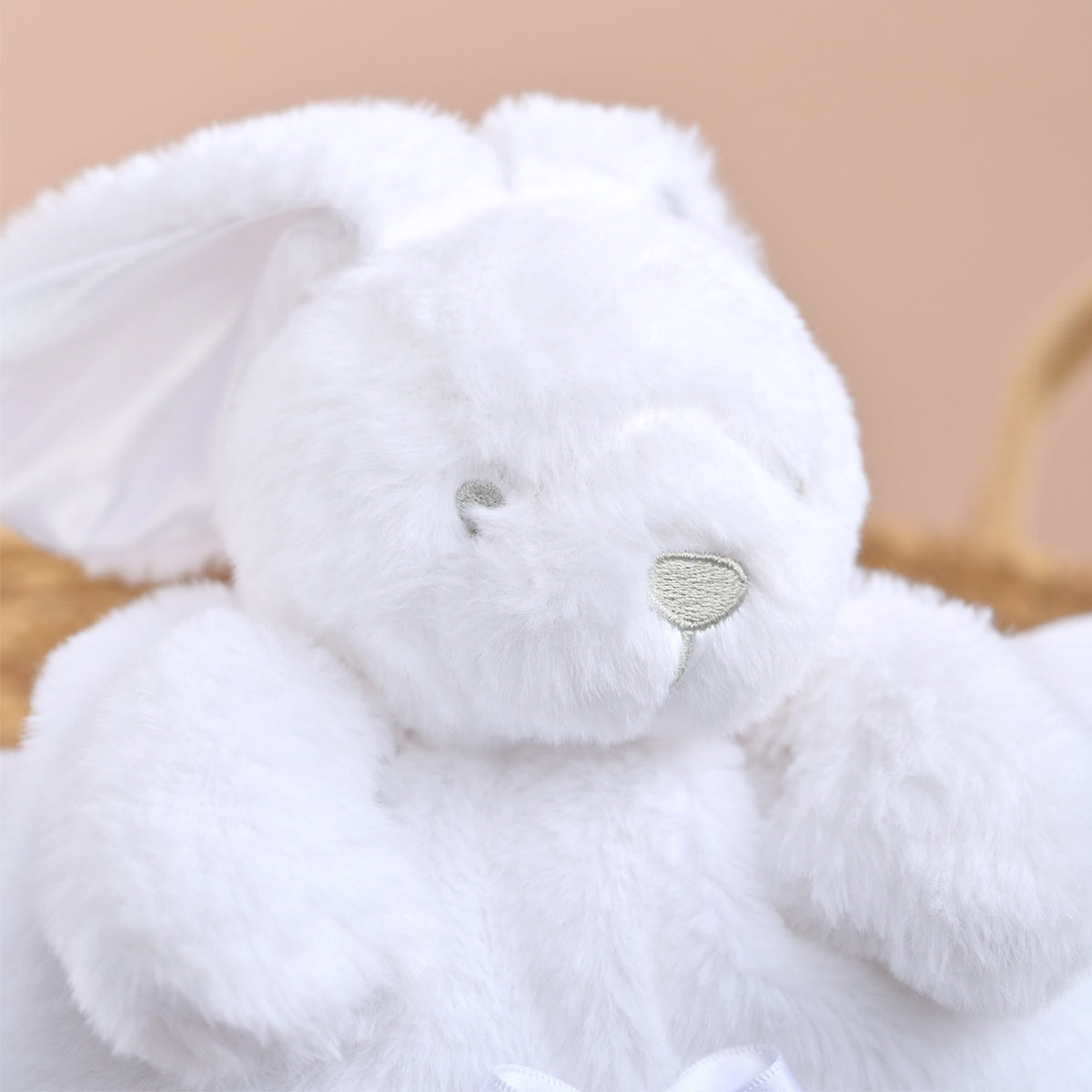 A close-up of a fluffy white plush bunny toy with long ears, small embroidered eyes, and a pale green nose, photographed against a soft beige background.