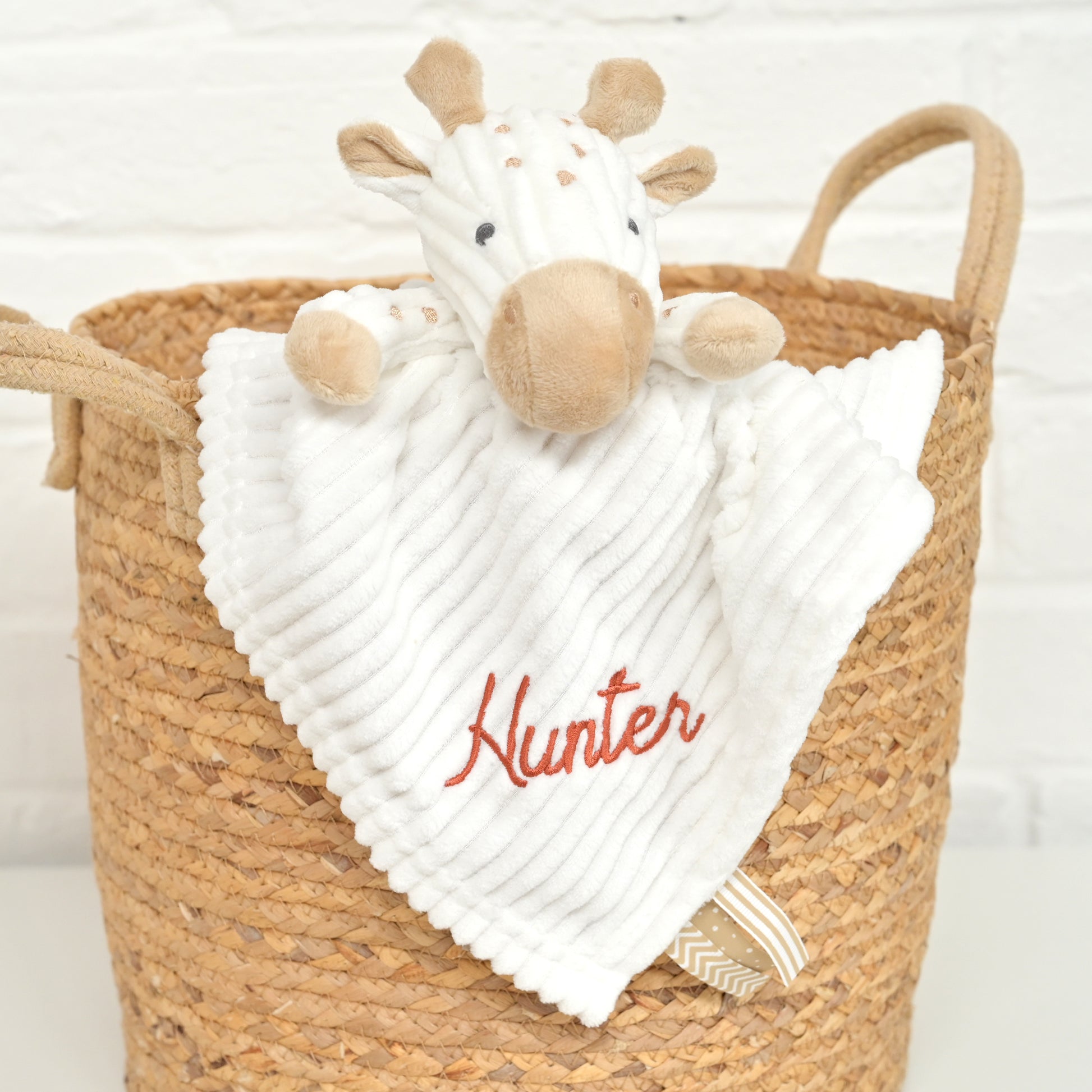 A white ribbed security blanket with a plush giraffe attached, personalized with the name 'Hunter' in red script, displayed inside a woven straw basket against a white brick wall.