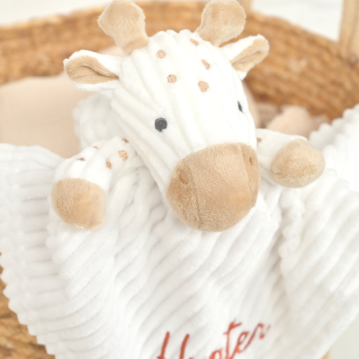 A close-up of a white plush giraffe lovey security blanket with a ribbed texture, featuring tan accents on the snout, ears, and spots, resting in a woven basket.
