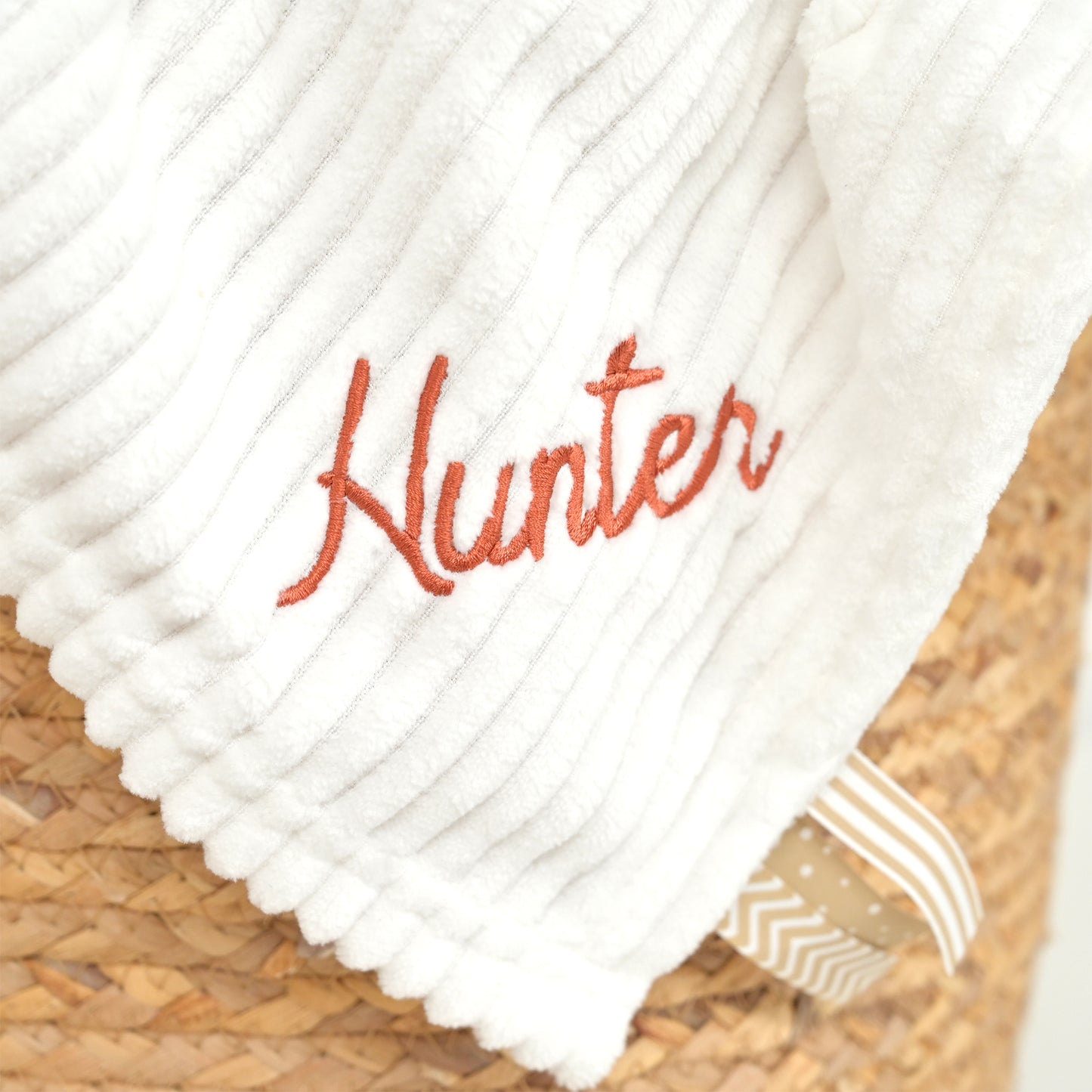 A close-up of a white ribbed fabric item, likely a blanket or throw, embroidered with the name 'Hunter' in orange script, resting on a woven straw surface.