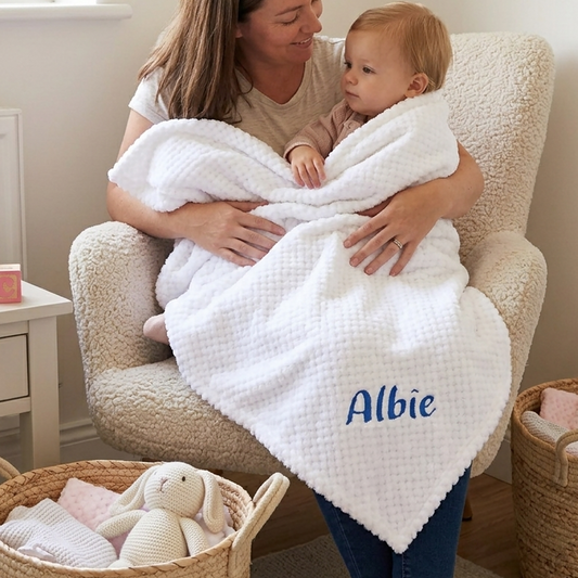 White waffle-knit baby blanket personalised with the name Albie embroidered in blue, held by his mother in a nursery chair.